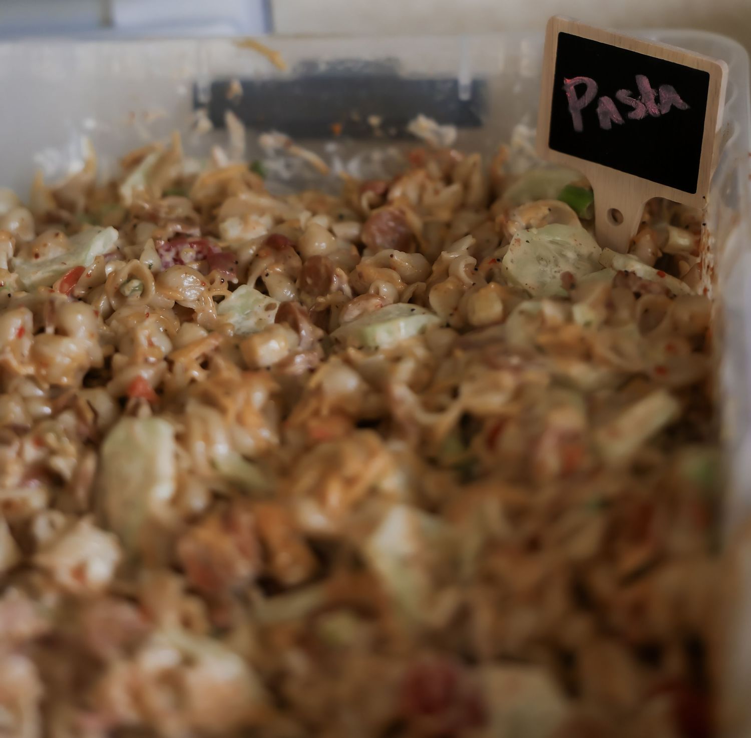 Large tray of creamy pasta salad with mixed vegetables labeled with a handwritten pasta sign.