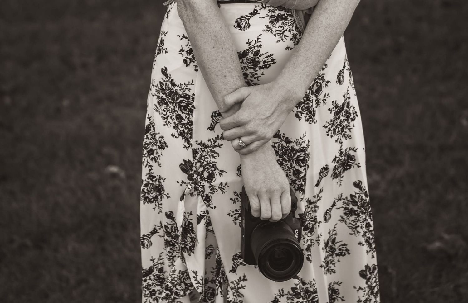 Black and white close-up of a woman in a floral dress holding a camera lens in both hands outdoors.