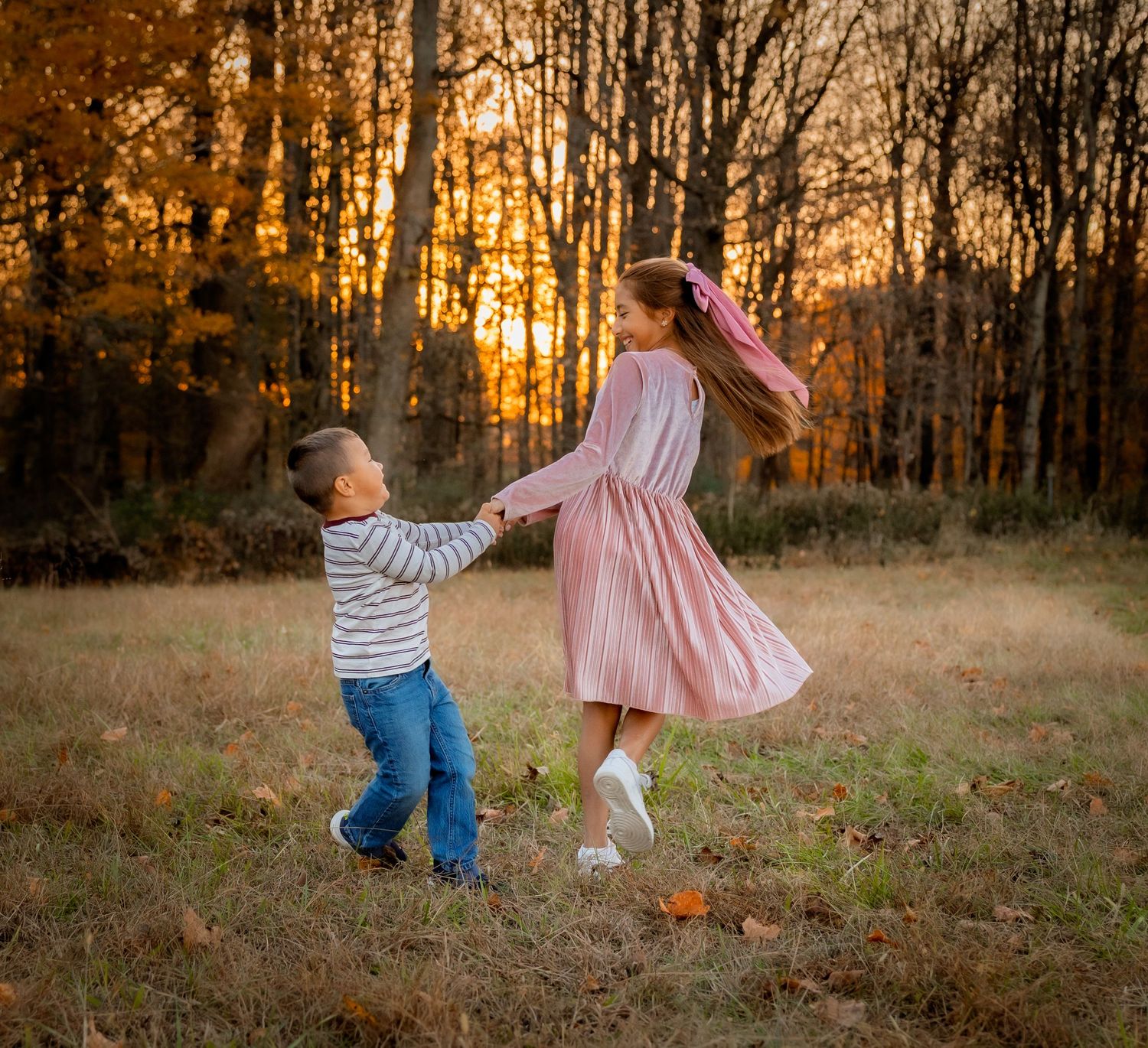 Two children hold hands and spin together in a golden autumn field at sunset, surrounded by fall foliage.