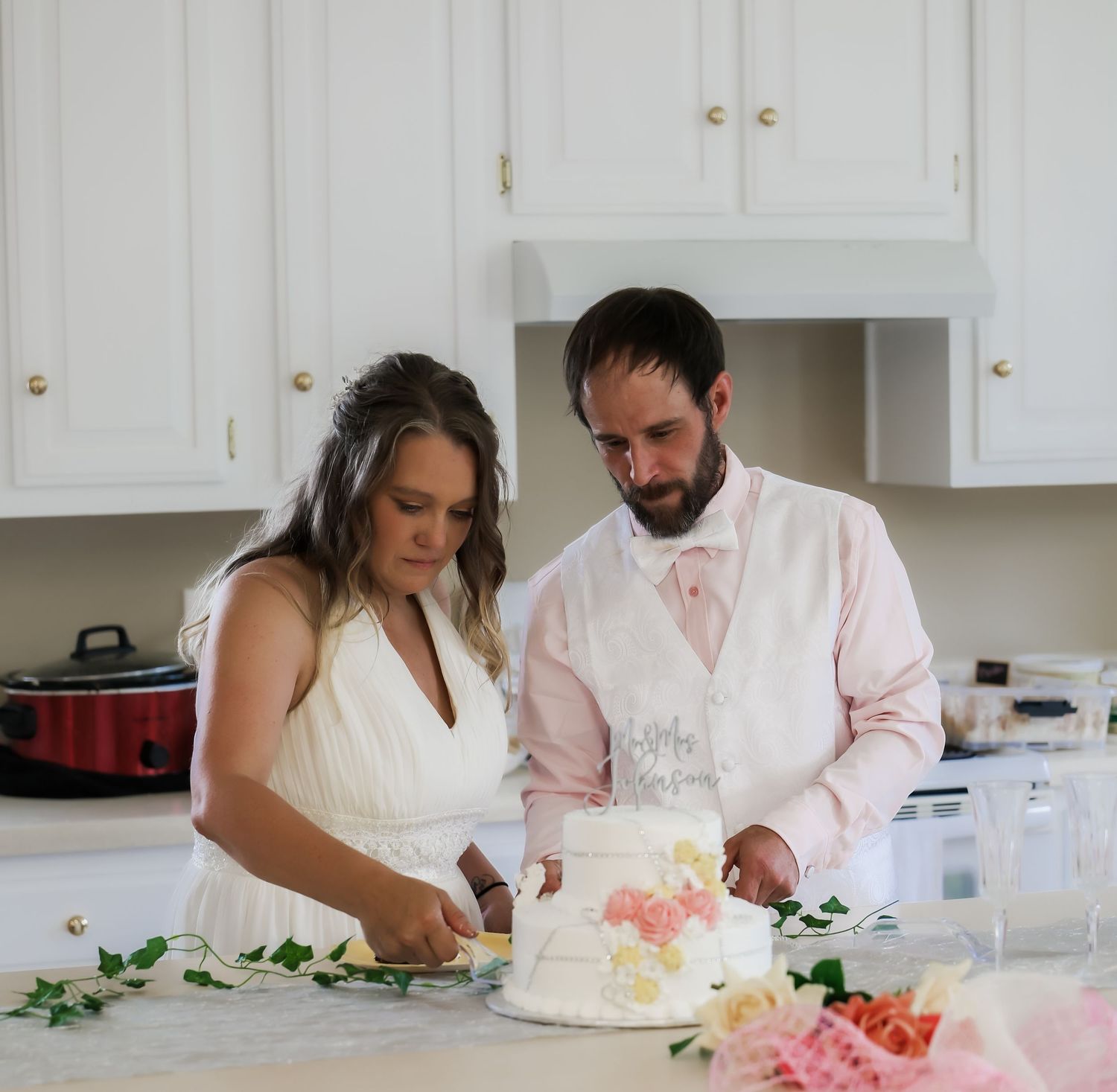Bride and groom cutting their wedding cake together in a white kitchen with floral decorations.