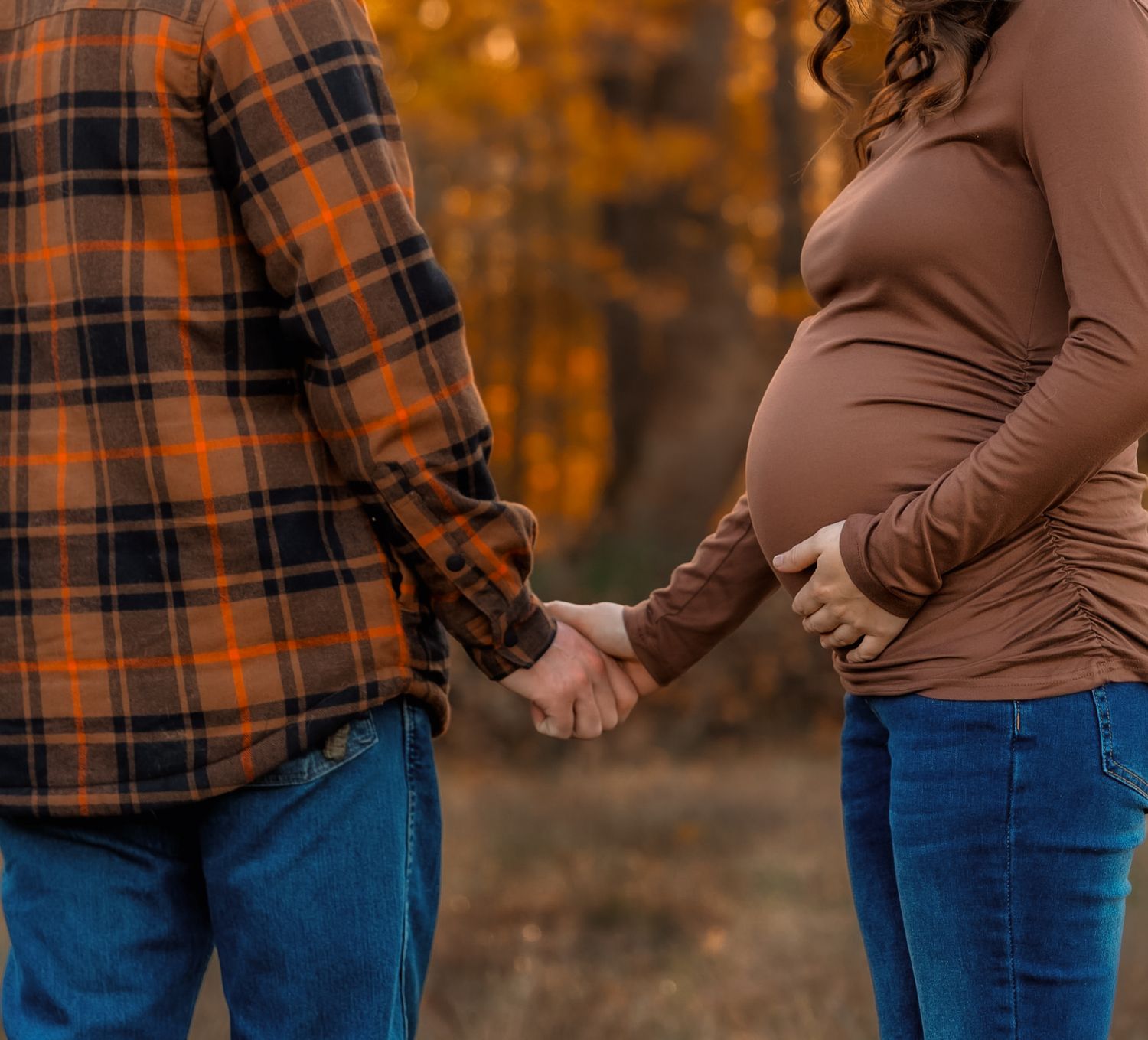 Pregnant couple holding hands outdoors in autumn, woman in brown top cradling baby bump, man in plaid shirt, fall foliage background.