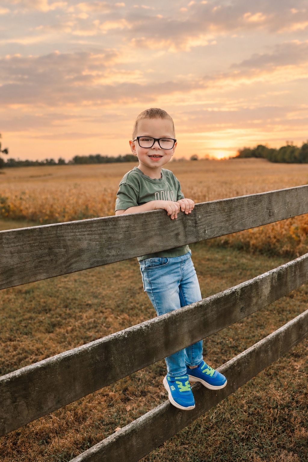 Young boy with glasses stands on wooden fence rail at sunset with golden field in background.