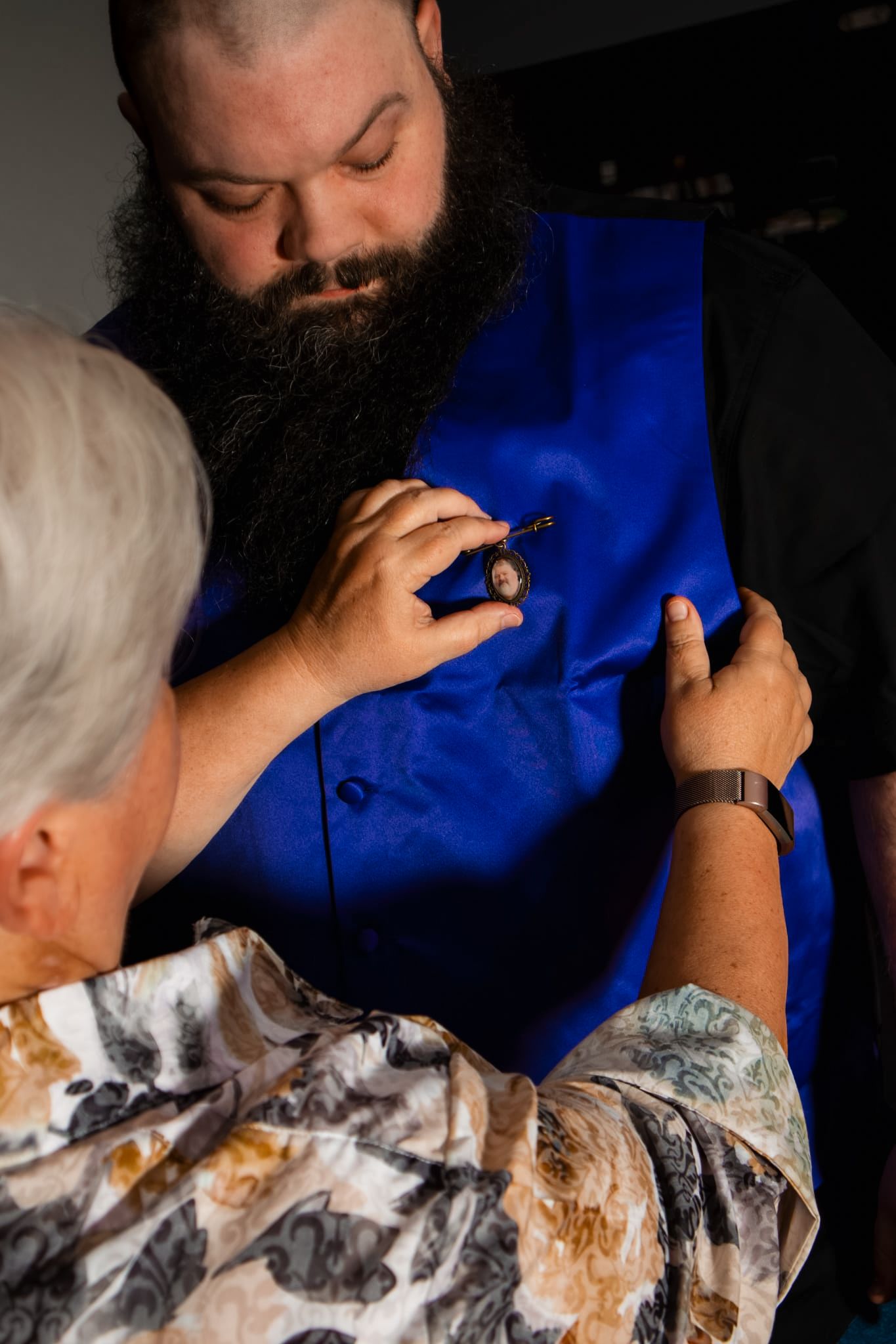 Older woman pinning a boutonniere on a bearded man wearing a royal blue vest at a formal event.