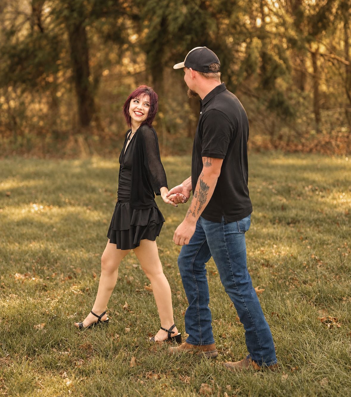 Couple holding hands and smiling in a park, woman in black dress with purple hair, man in black polo and jeans.