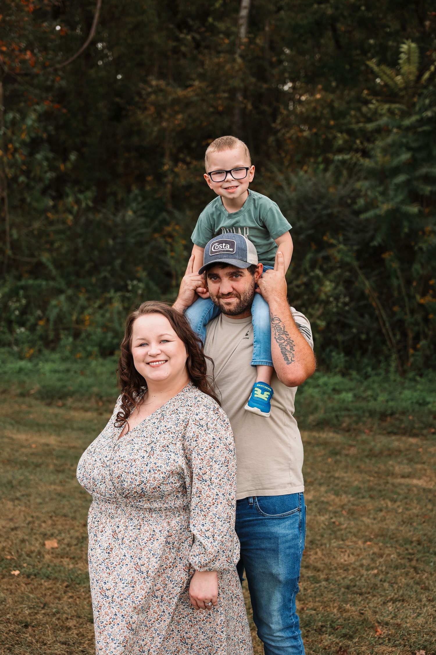 A smiling family of three poses outdoors, with a young boy sitting on his fathers shoulders, surrounded by lush green trees.