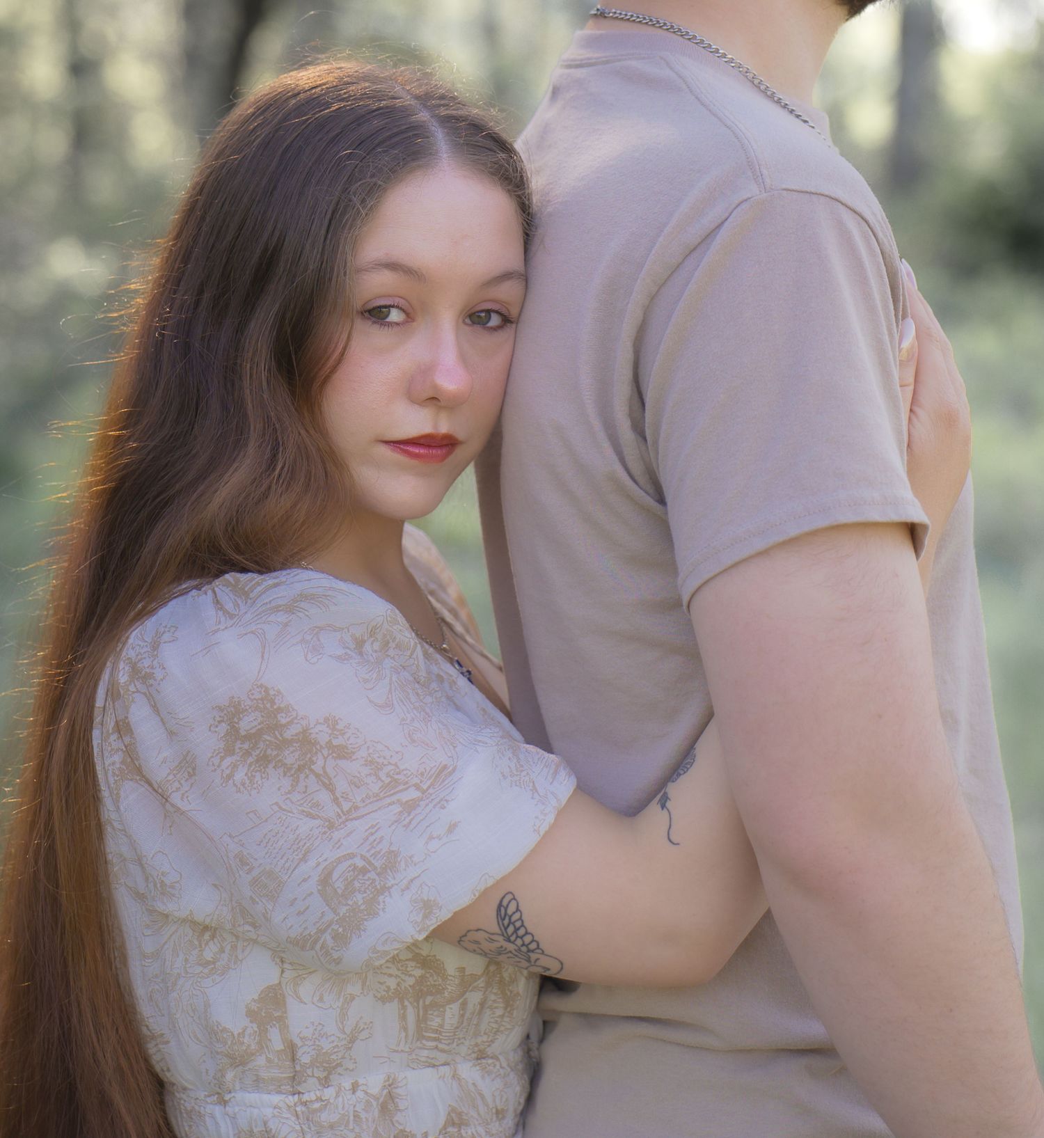 Young woman with long hair in a floral dress embraces a man from behind in a softly lit outdoor setting.