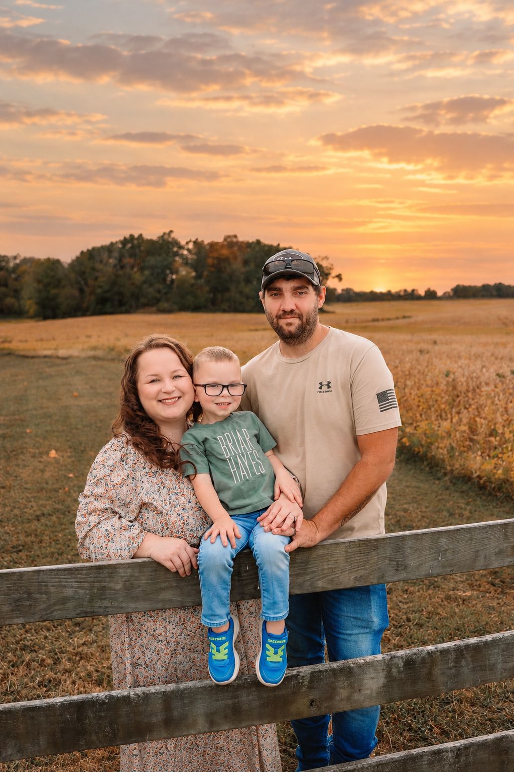 Family of three posing by a wooden fence at golden hour sunset with open fields and colorful sky in background.