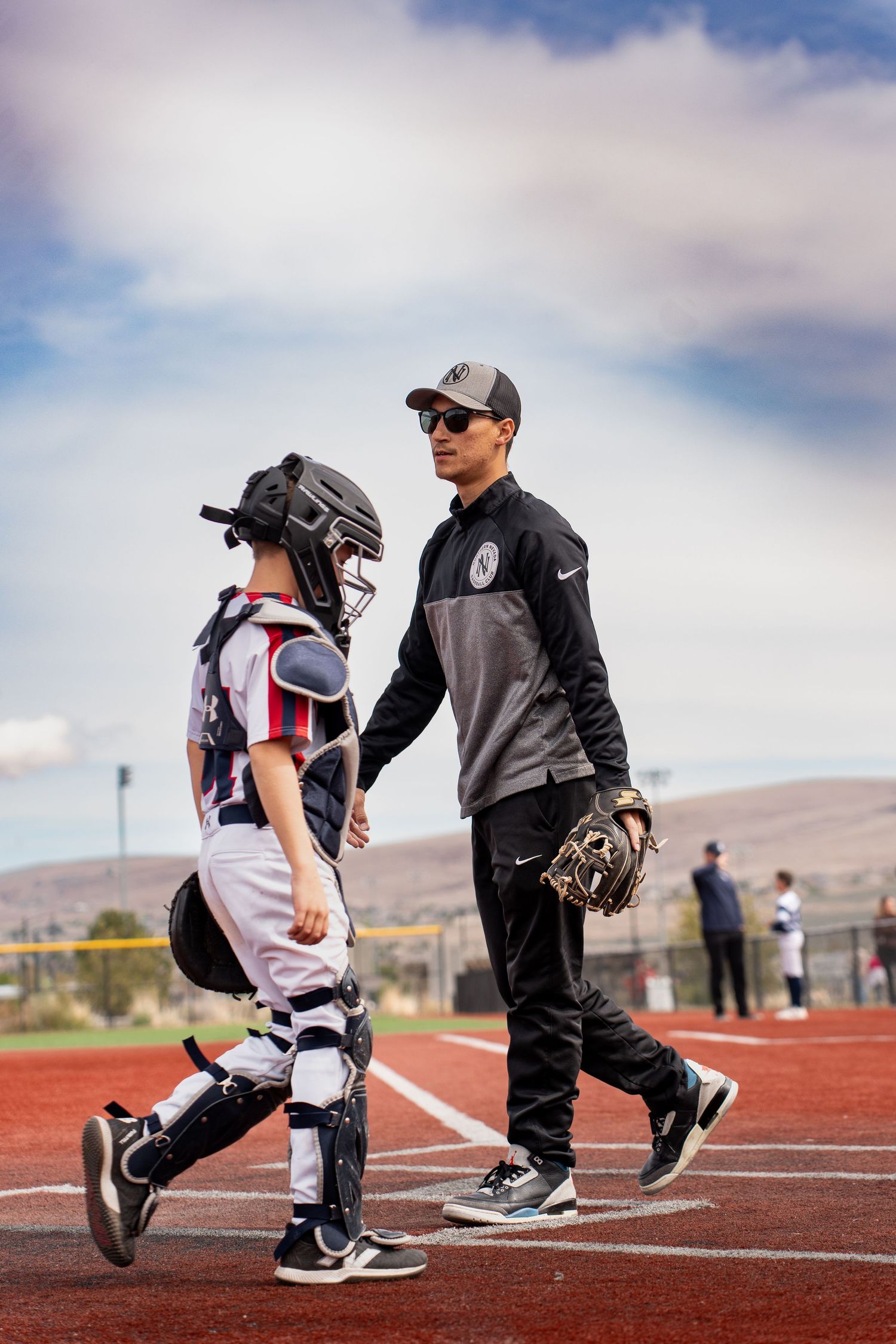 Baseball coach walking with young catcher in full gear on an outdoor baseball field.
