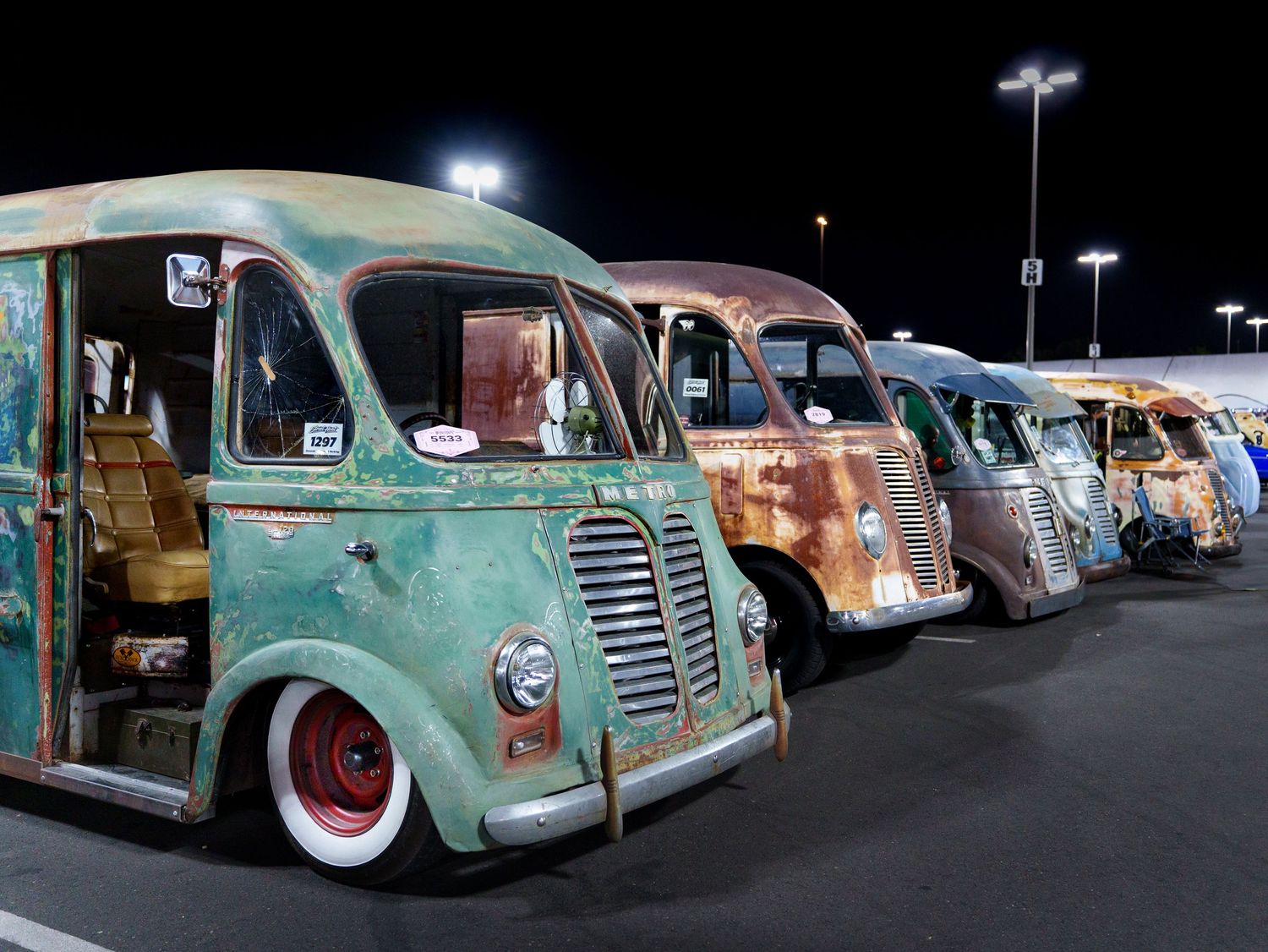 Row of vintage rusty patina delivery vans displayed at a nighttime classic car show.