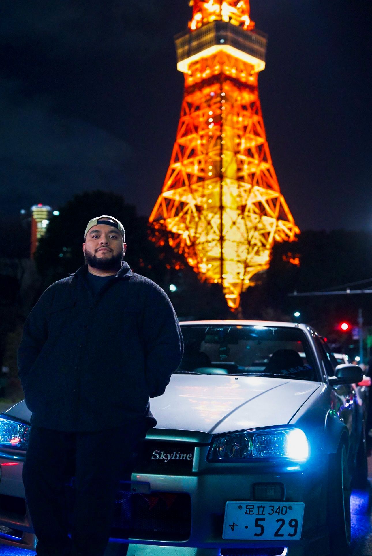 Man posing in front of illuminated Tokyo Tower at night beside a Nissan Skyline sports car.