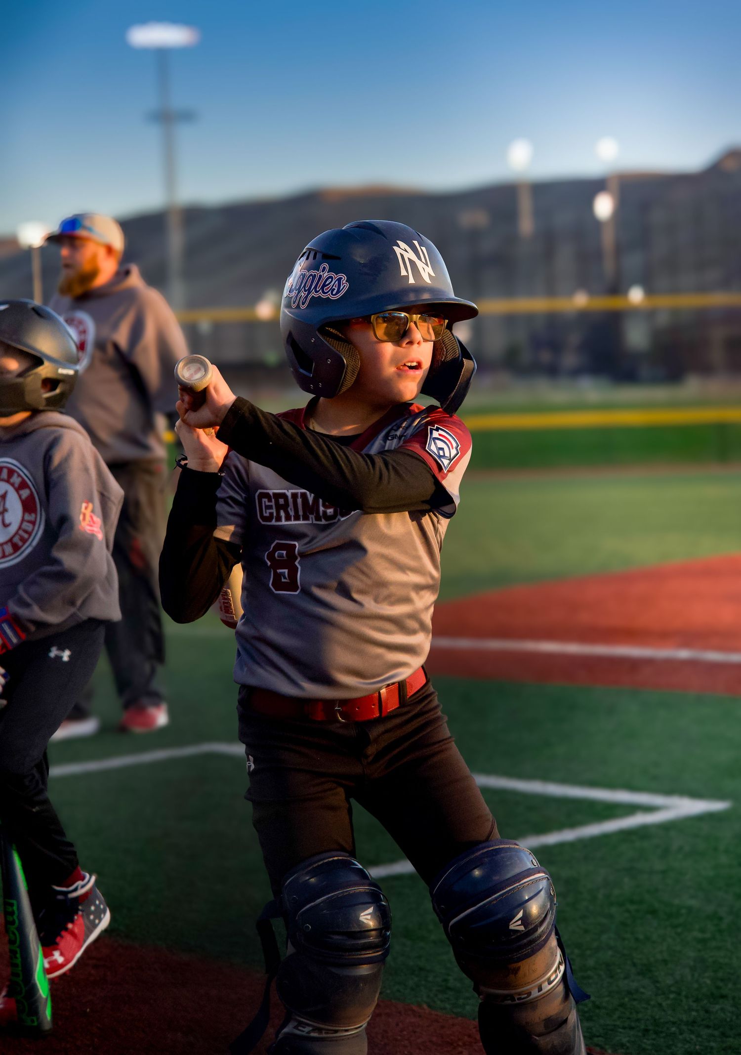 Young baseball player in Crimson jersey number 8 holds bat in batting stance wearing full catcher gear.