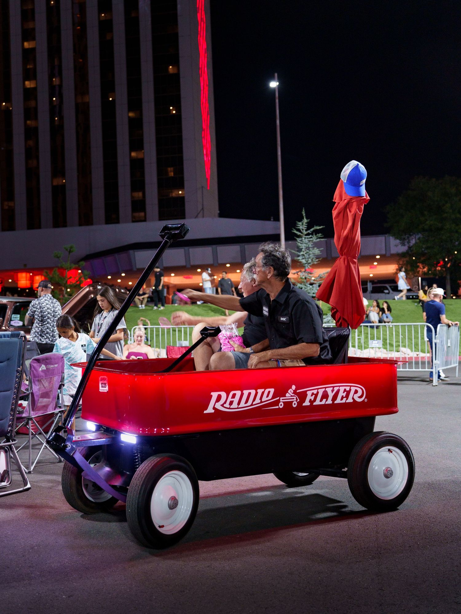 Man riding a giant red Radio Flyer wagon vehicle at an outdoor nighttime event.