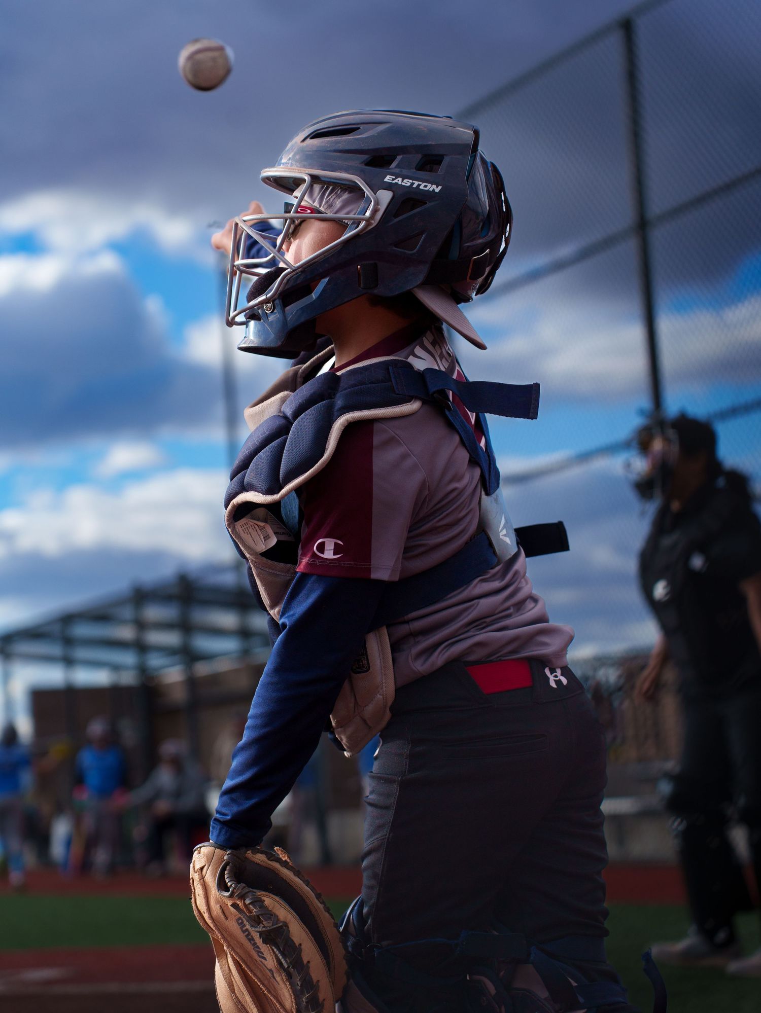 Youth baseball catcher in Easton helmet and full gear tracks incoming ball against dramatic cloudy sky backdrop.