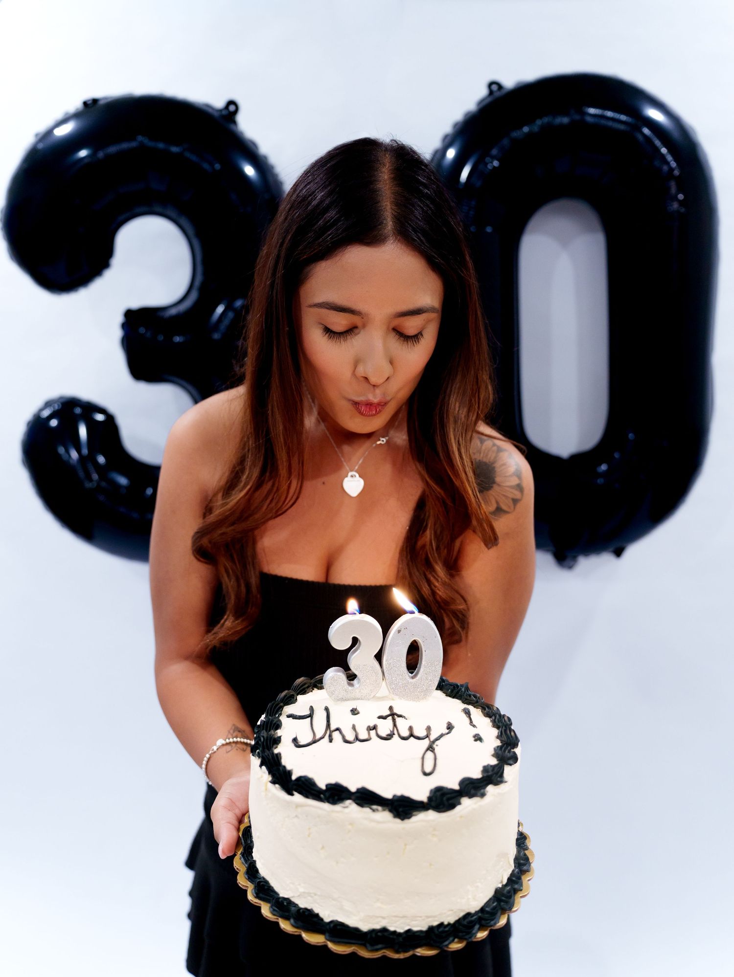 A woman blows out a candle on a white 30th birthday cake with black number balloons behind her.