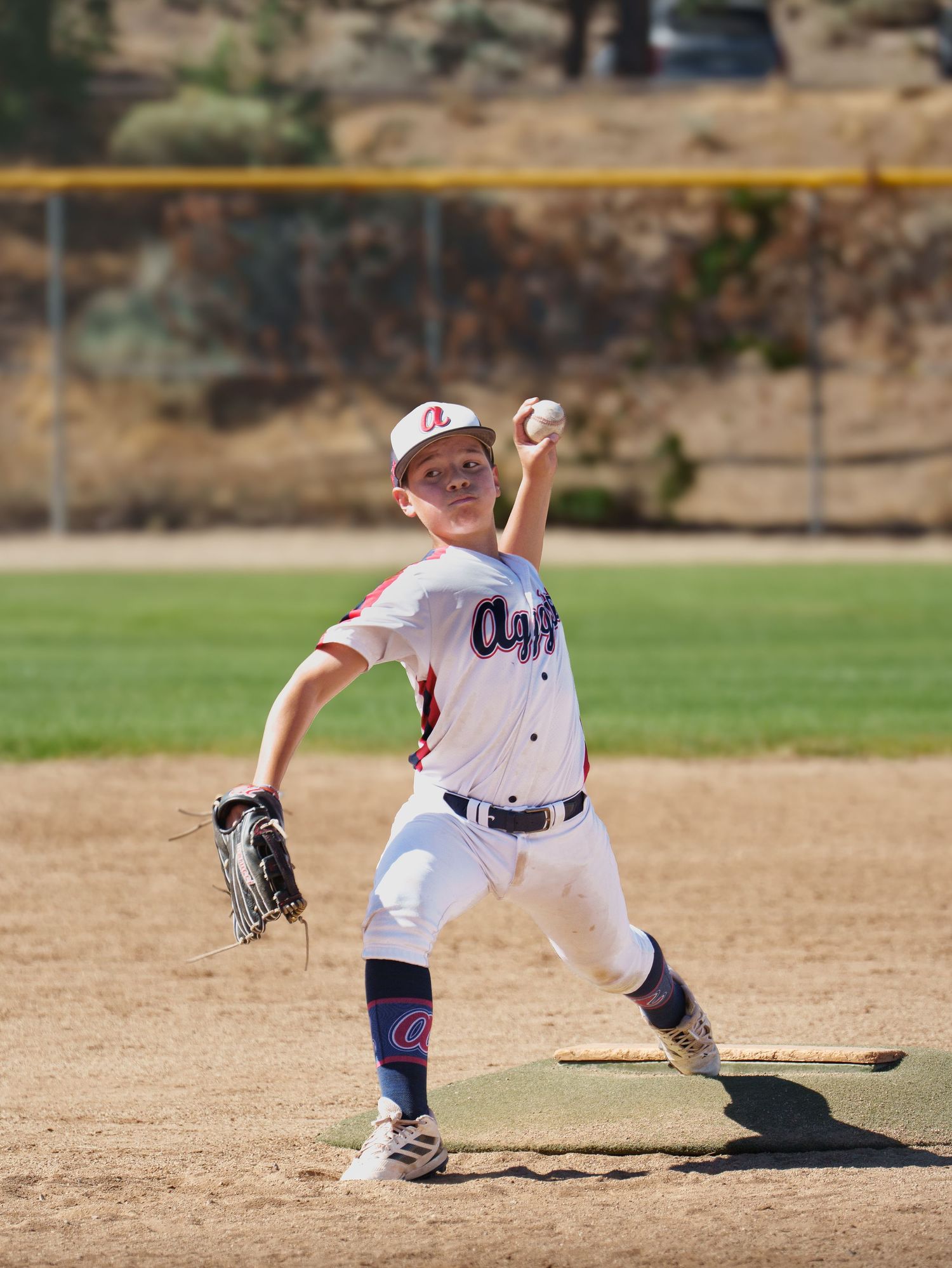Young pitcher in white Aggies uniform throwing ball on sunny outdoor baseball diamond.