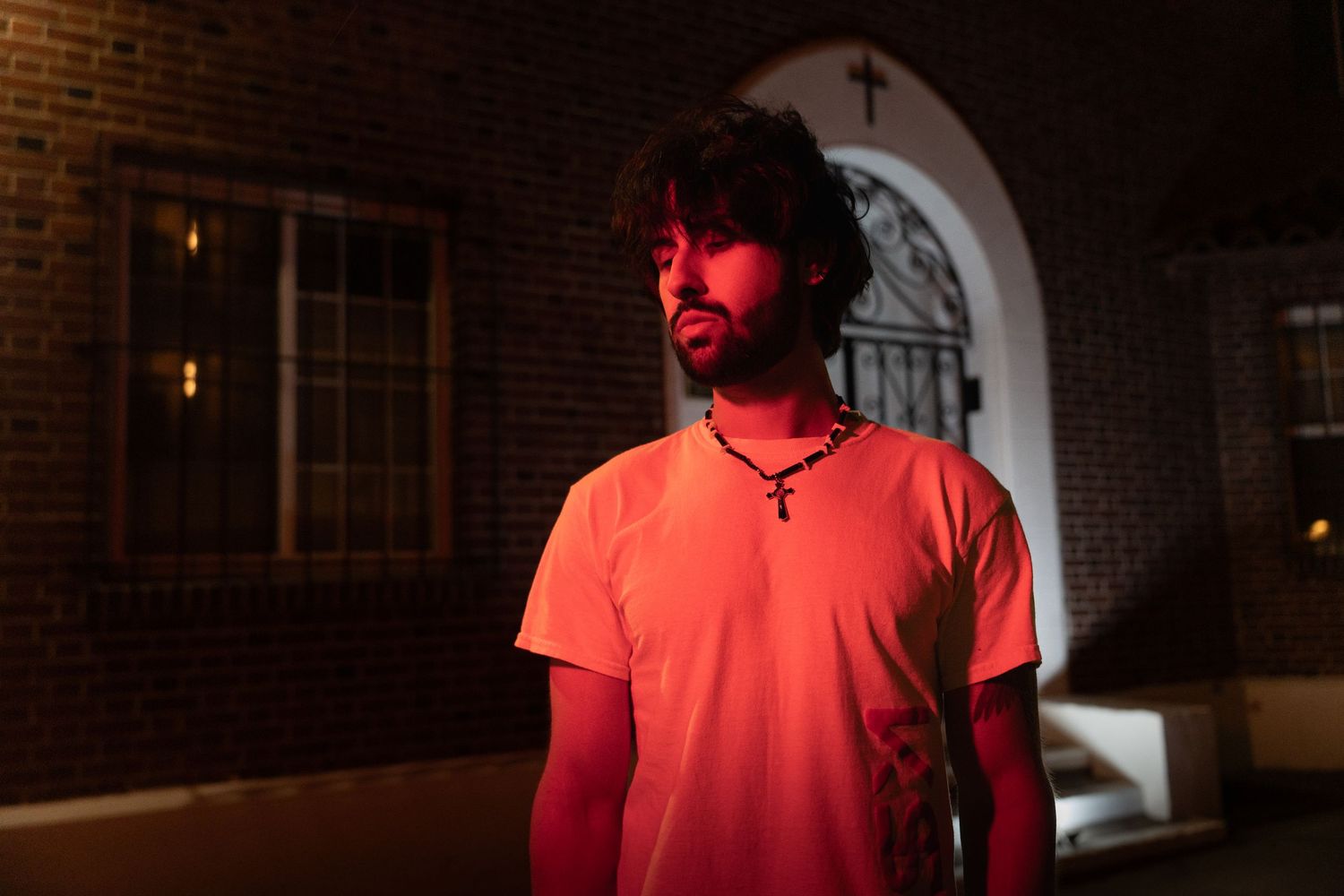 Bearded man with cross necklace in white t-shirt stands in red light by brick church at night.