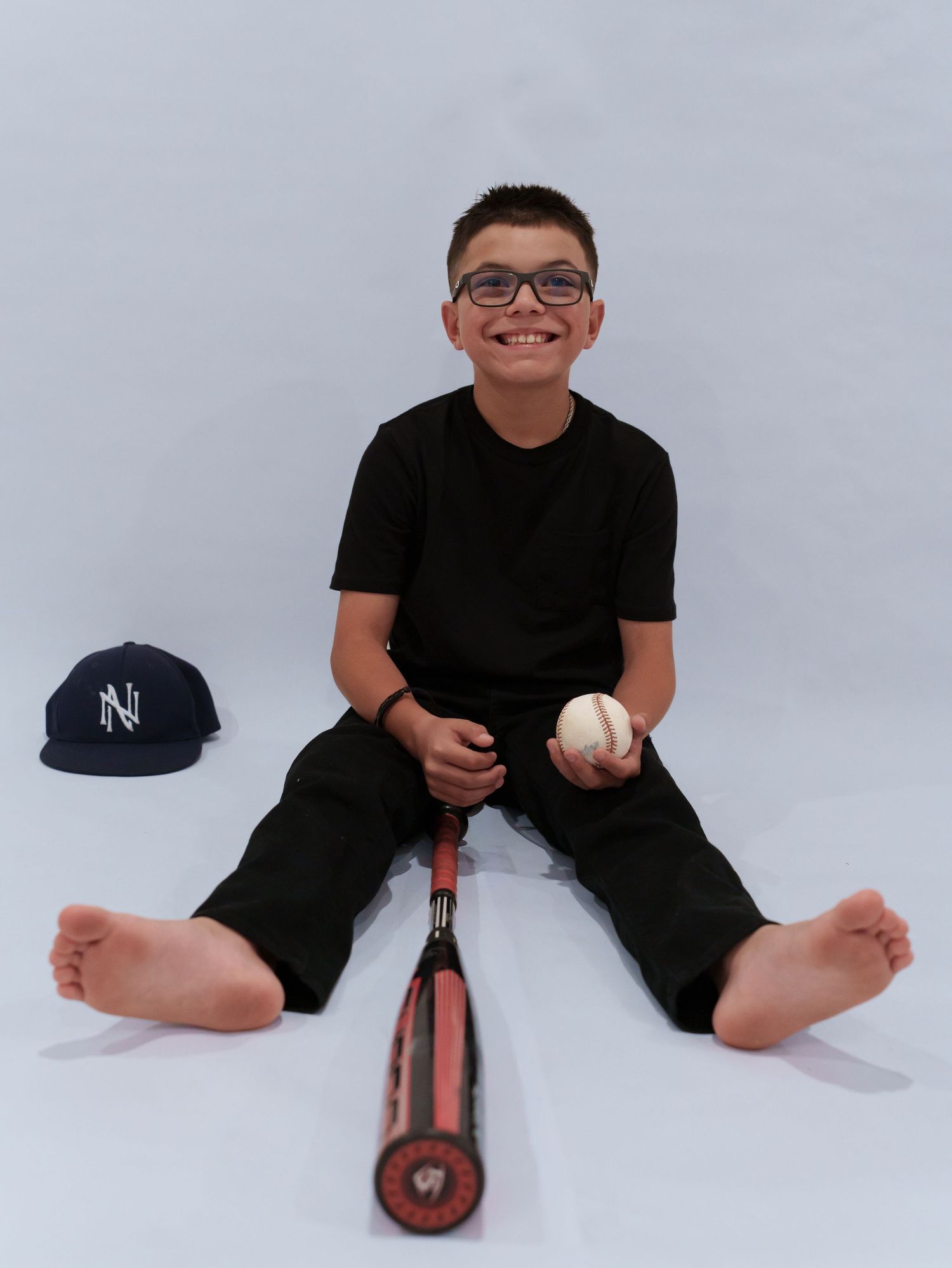 Smiling boy in black outfit sits barefoot with baseball bat, glove, and NY cap on white background.