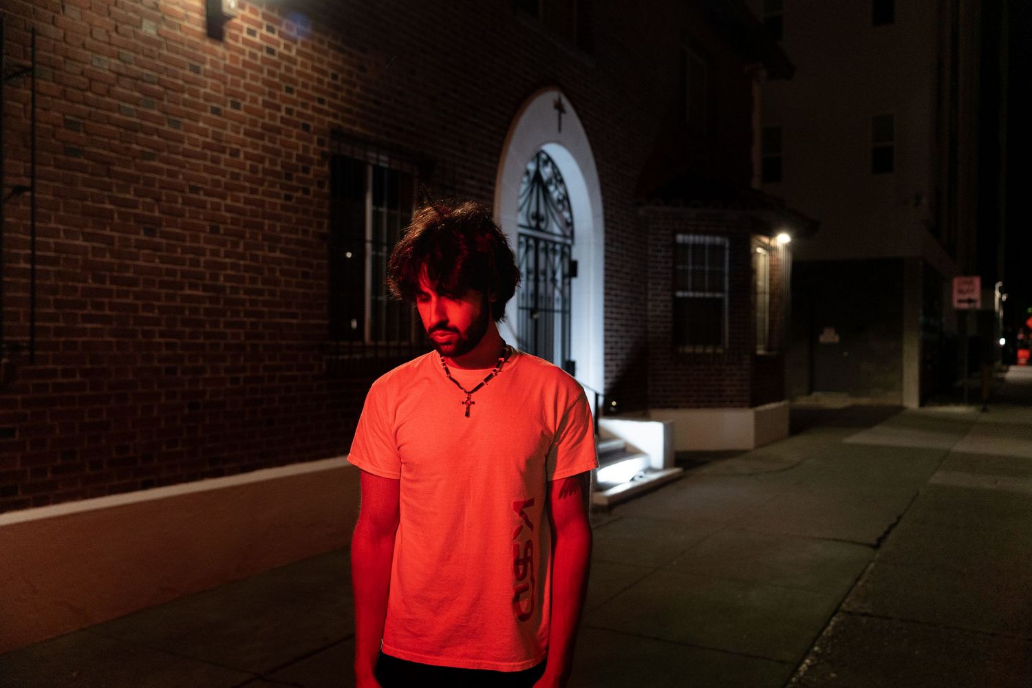 Tattooed man in white t-shirt looks down on sidewalk outside brick building at night.