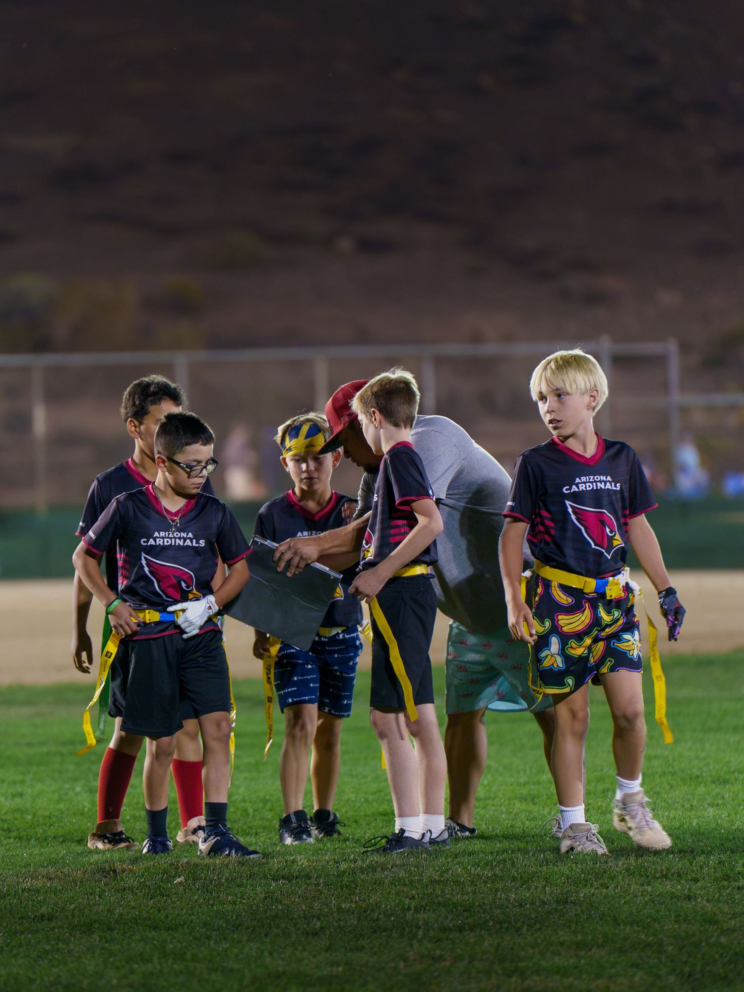 Youth flag football players in Arizona Cardinals jerseys receiving coaching during an evening game under lights.