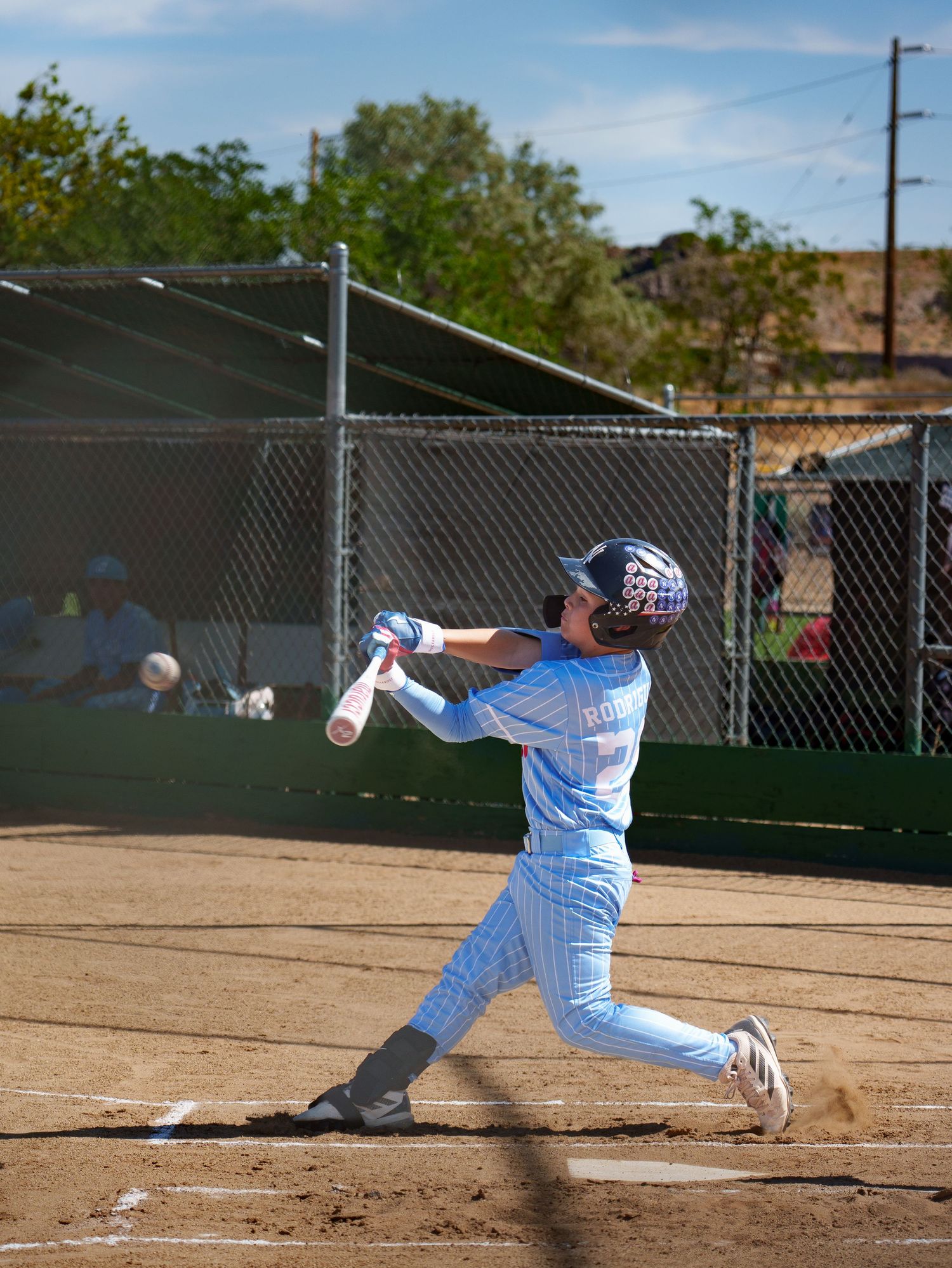 Youth baseball player swings bat during game wearing light blue pinstripe uniform at outdoor baseball field.