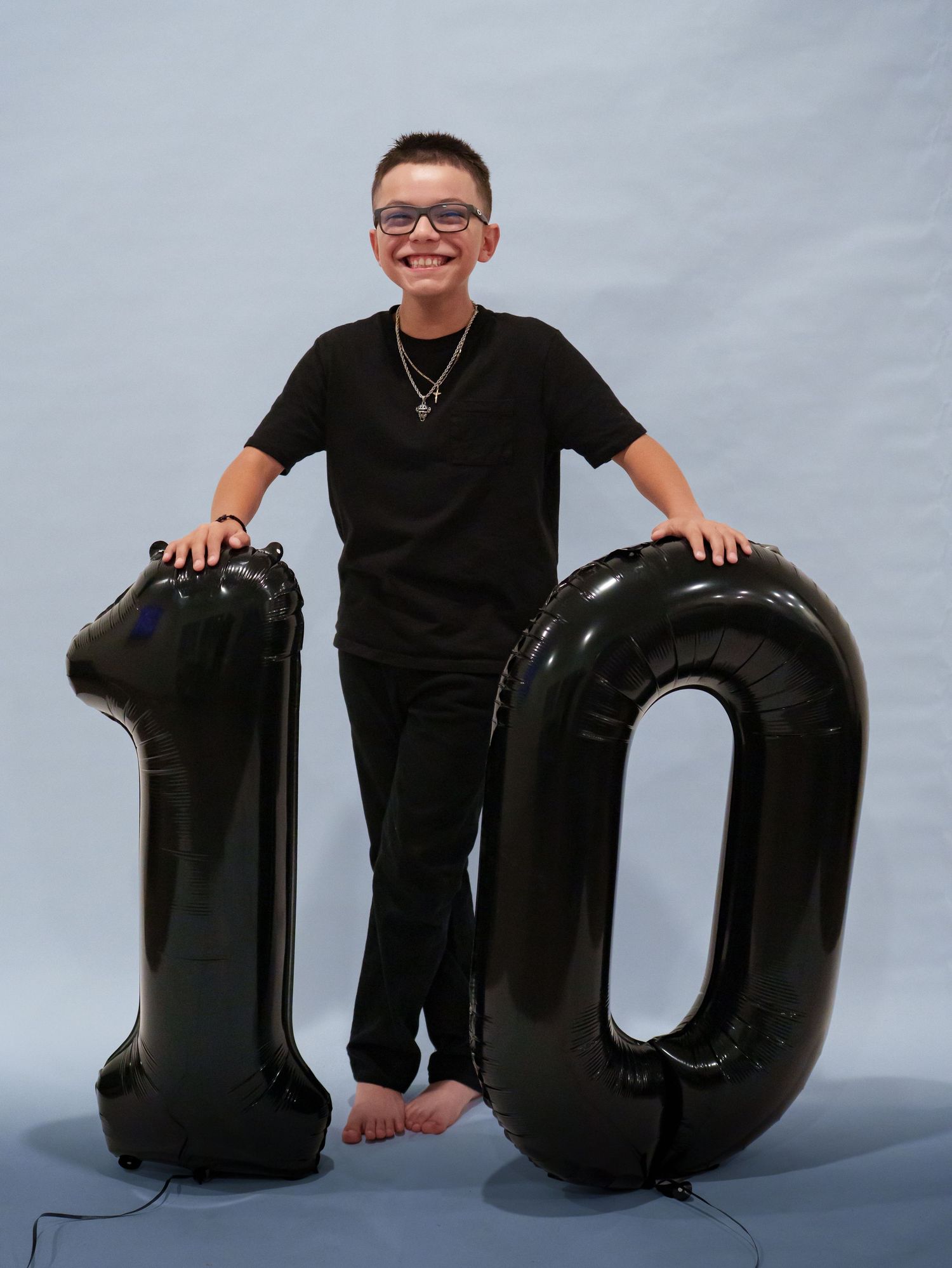 A smiling boy in all black stands between large black number 10 balloons celebrating his 10th birthday.
