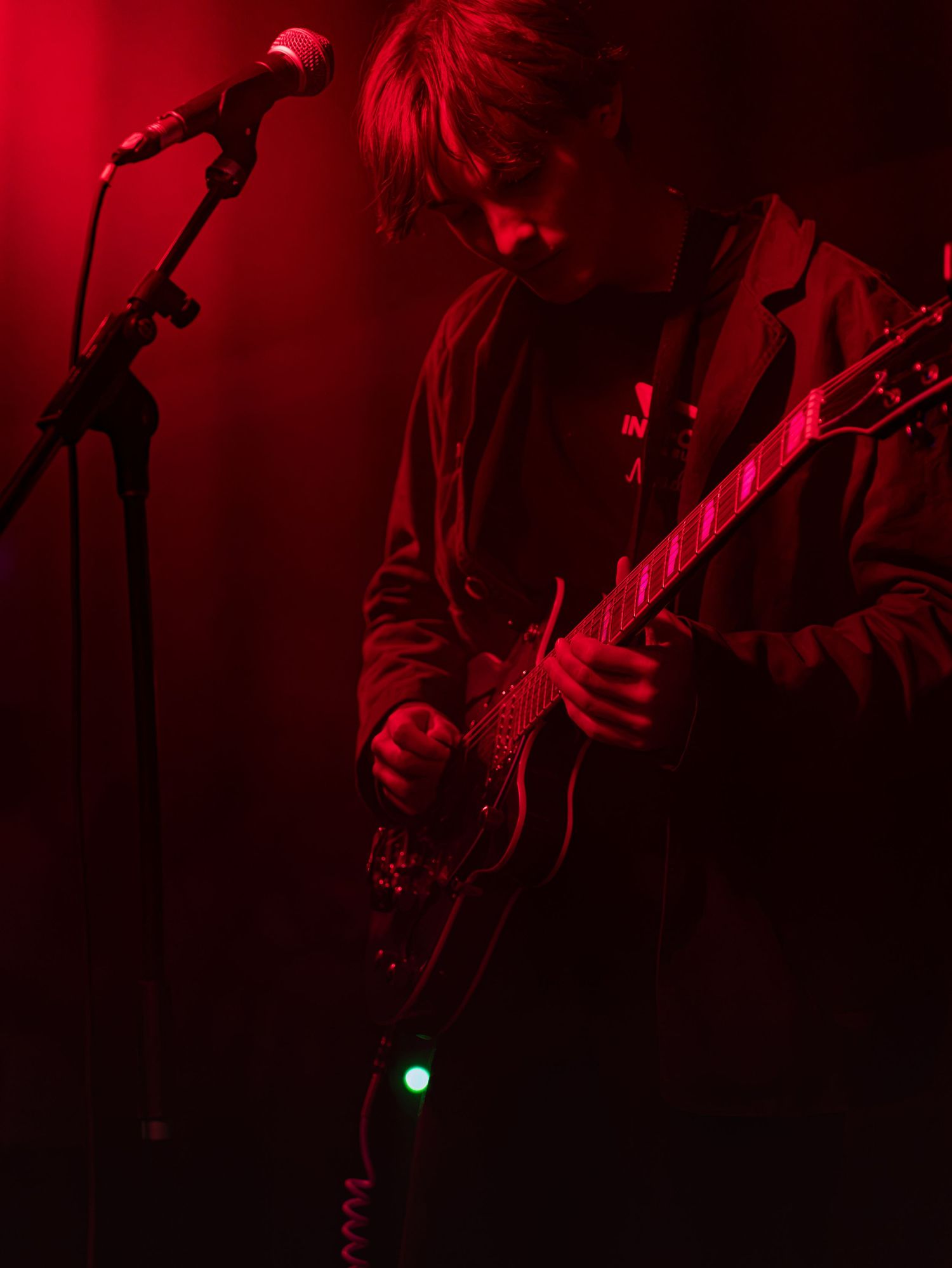 A guitarist plays an electric guitar on stage under moody red concert lighting.