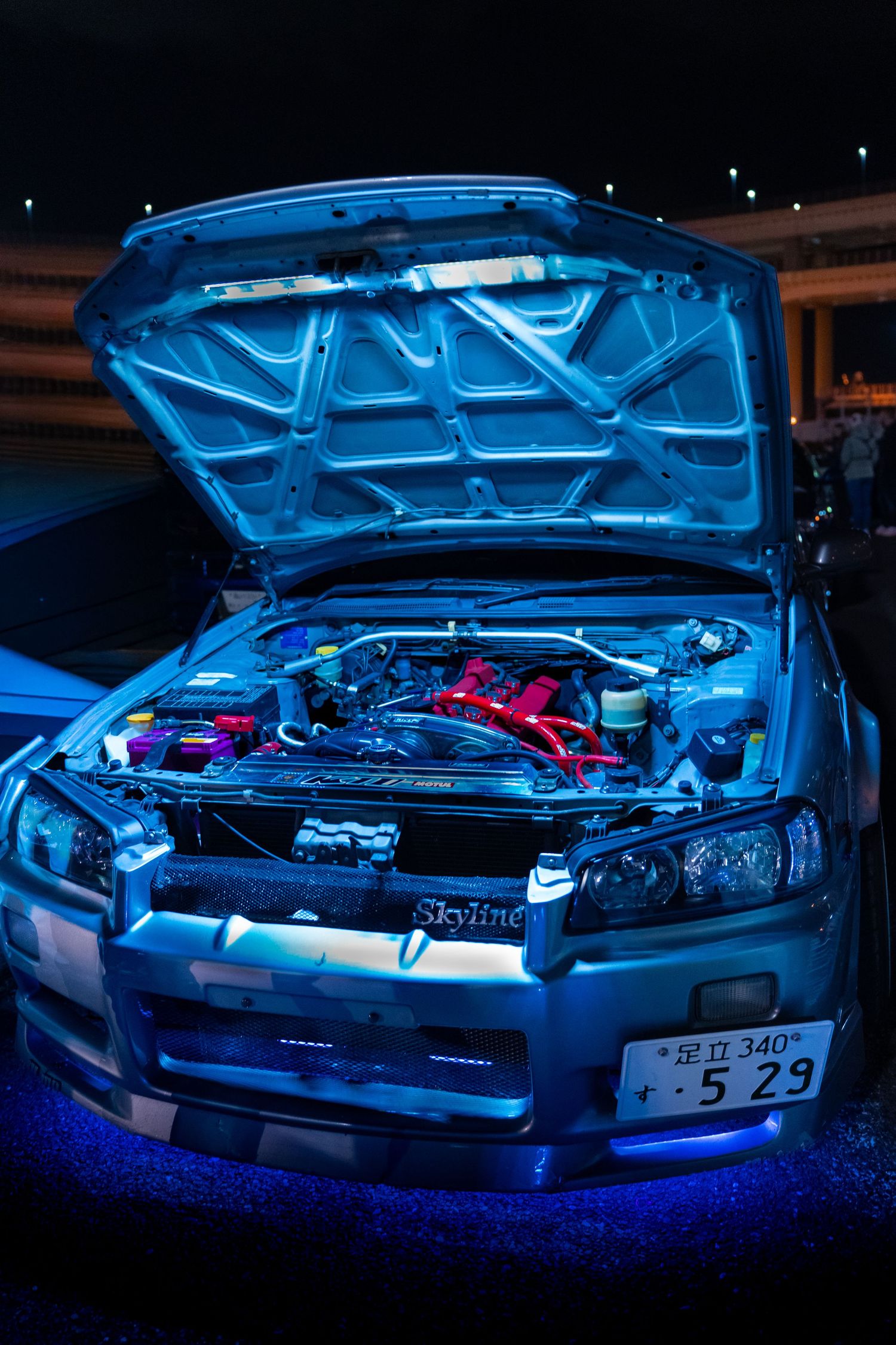 Nissan Skyline R34 with hood open revealing engine bay illuminated by blue underglow lighting at night.