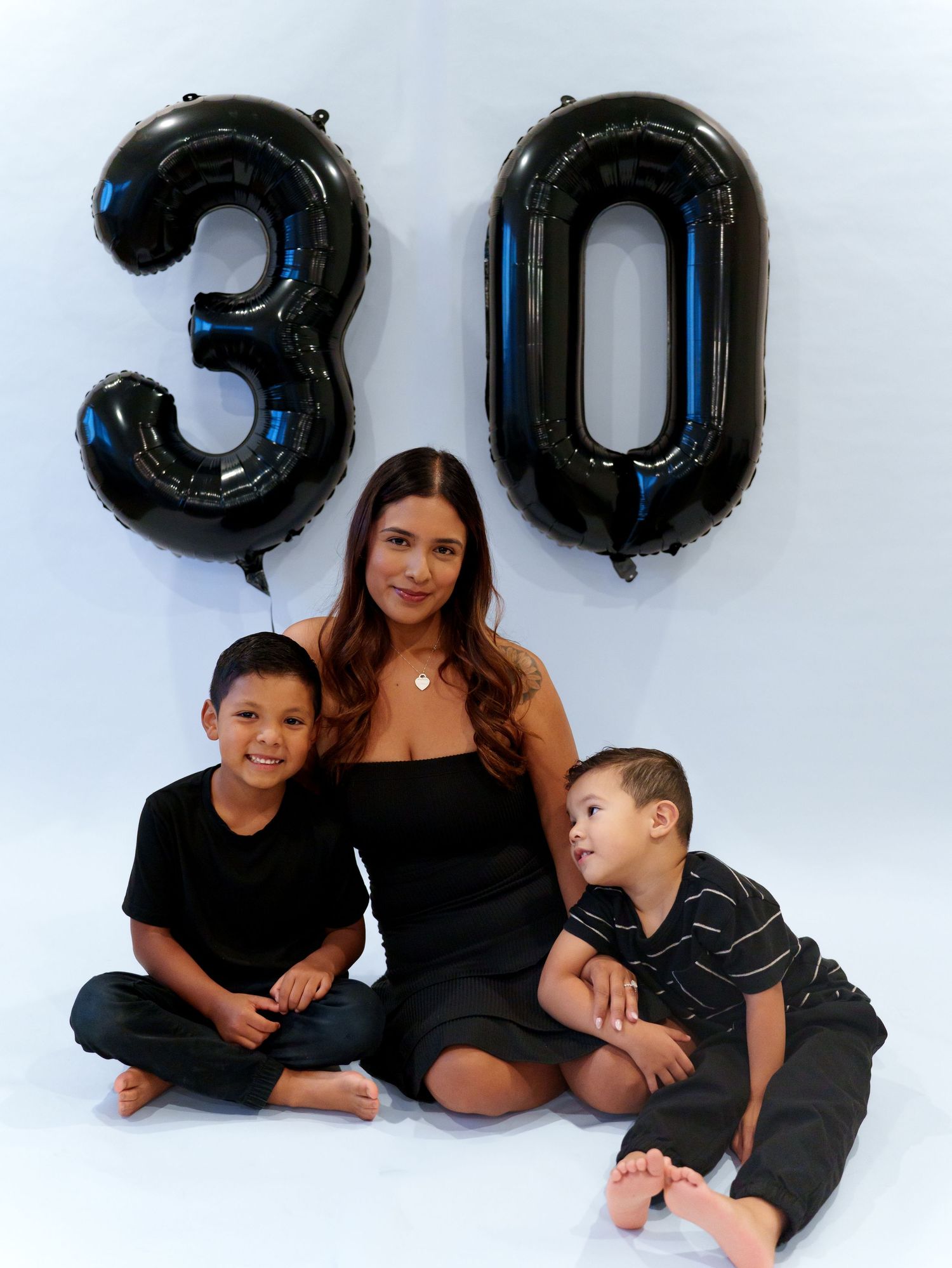 Woman celebrating 30th birthday sitting with two young boys beside large black number balloons.
