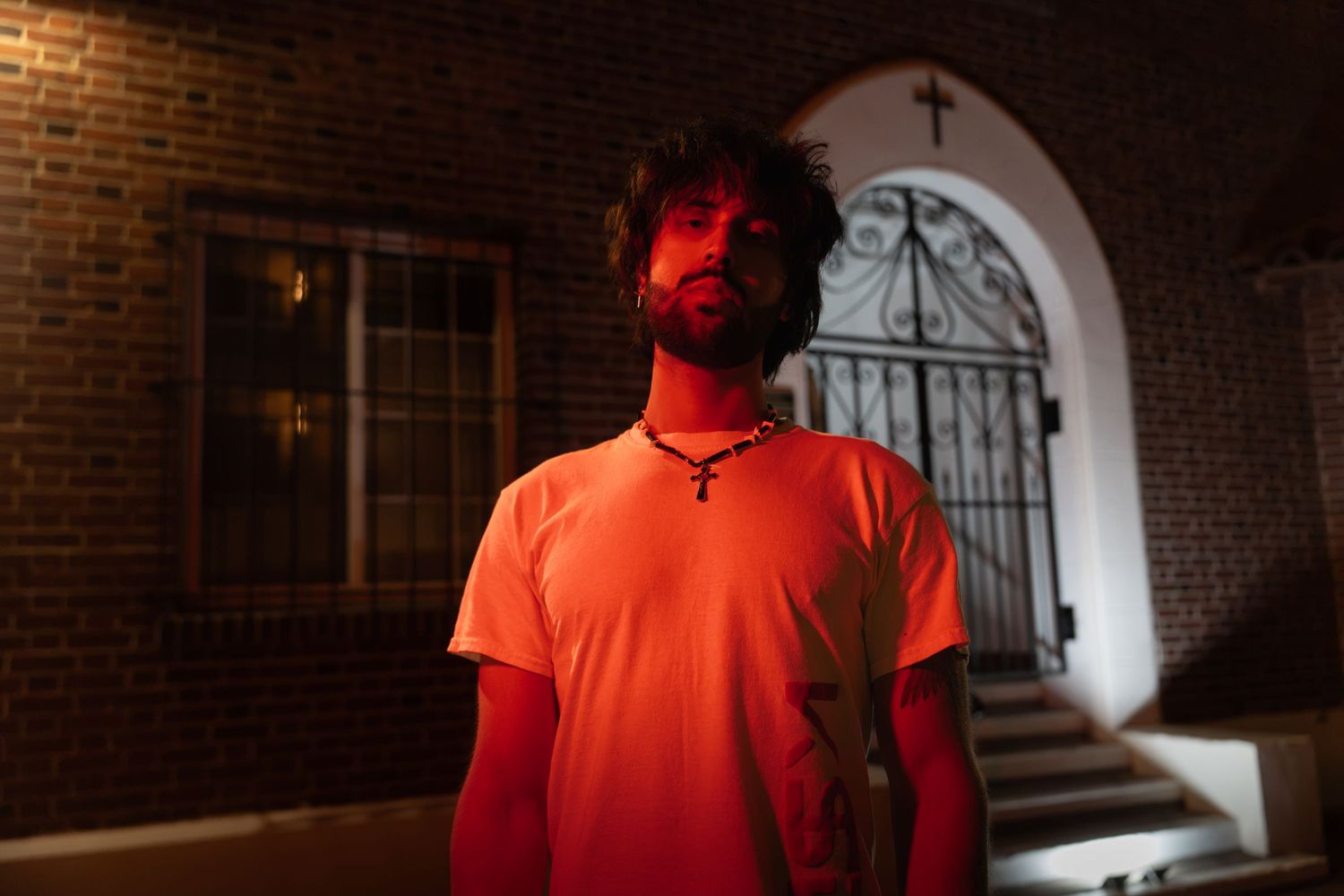 Bearded man in orange t-shirt stands in red light before brick building with cross at night.