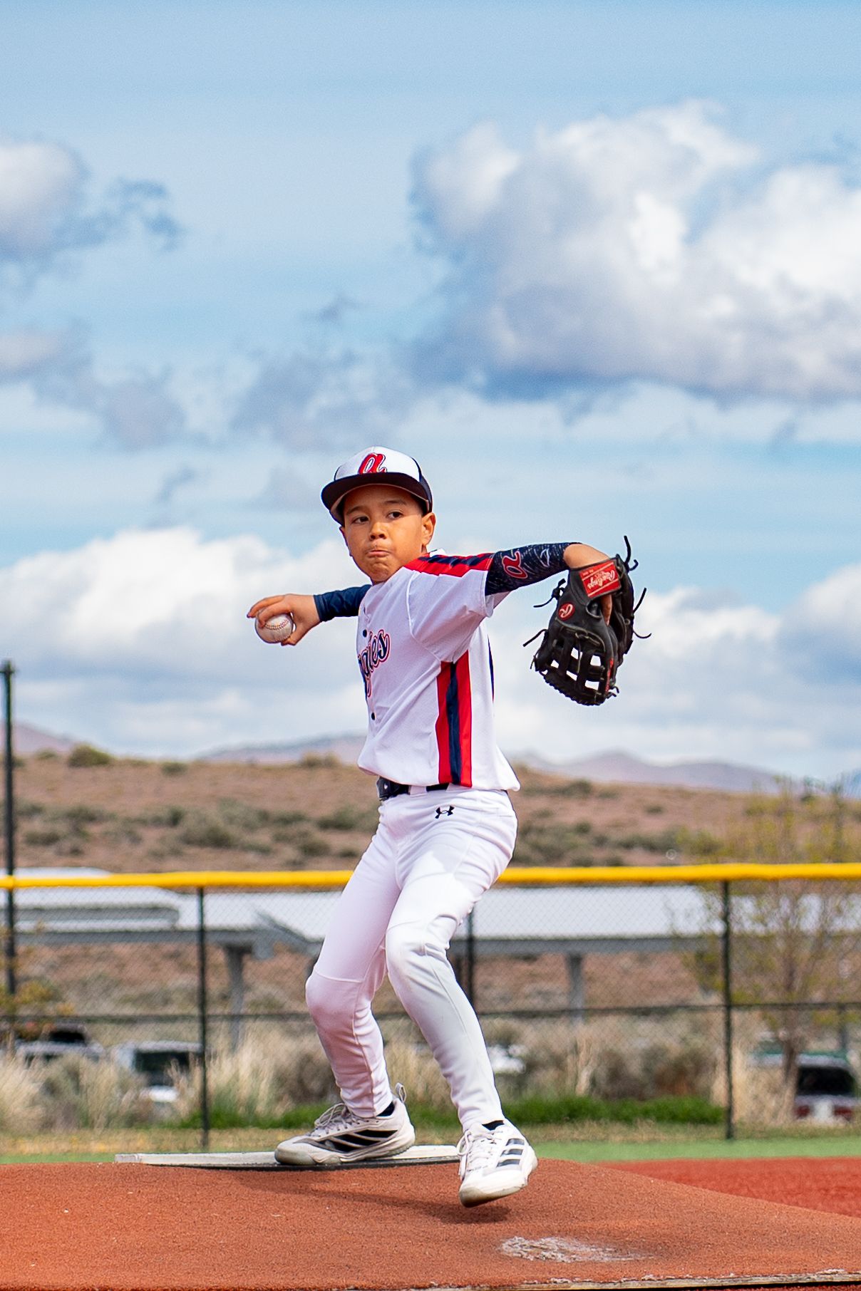 Young baseball pitcher winds up to throw on mound wearing white uniform with red and blue details outdoors.
