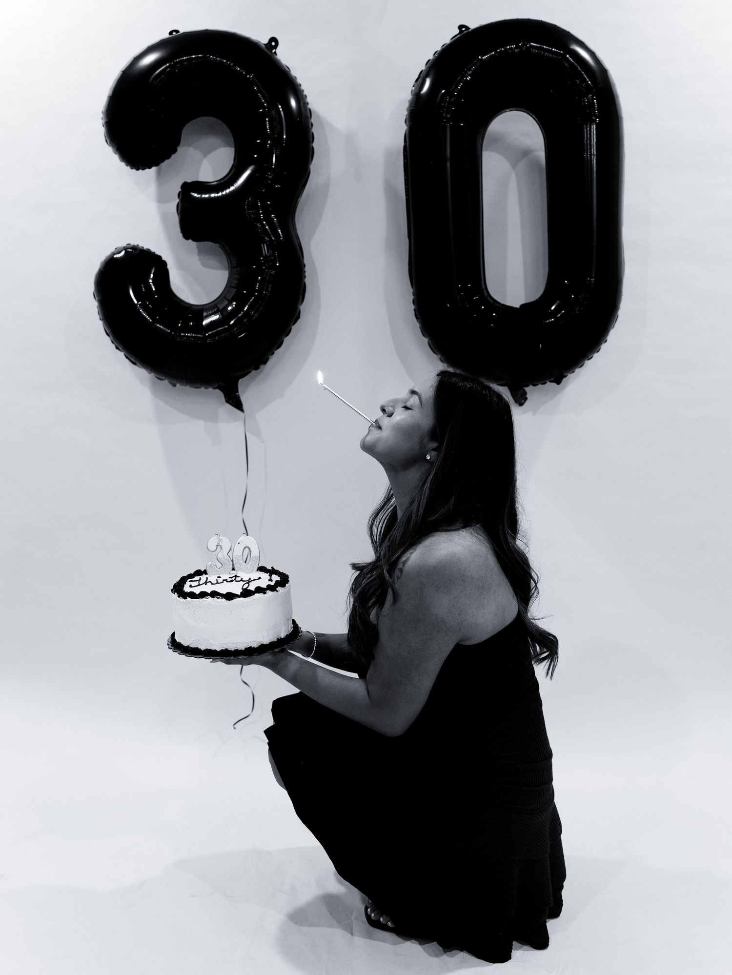Black and white photo of a woman kneeling and blowing out candles on a cake with number 30 balloons.