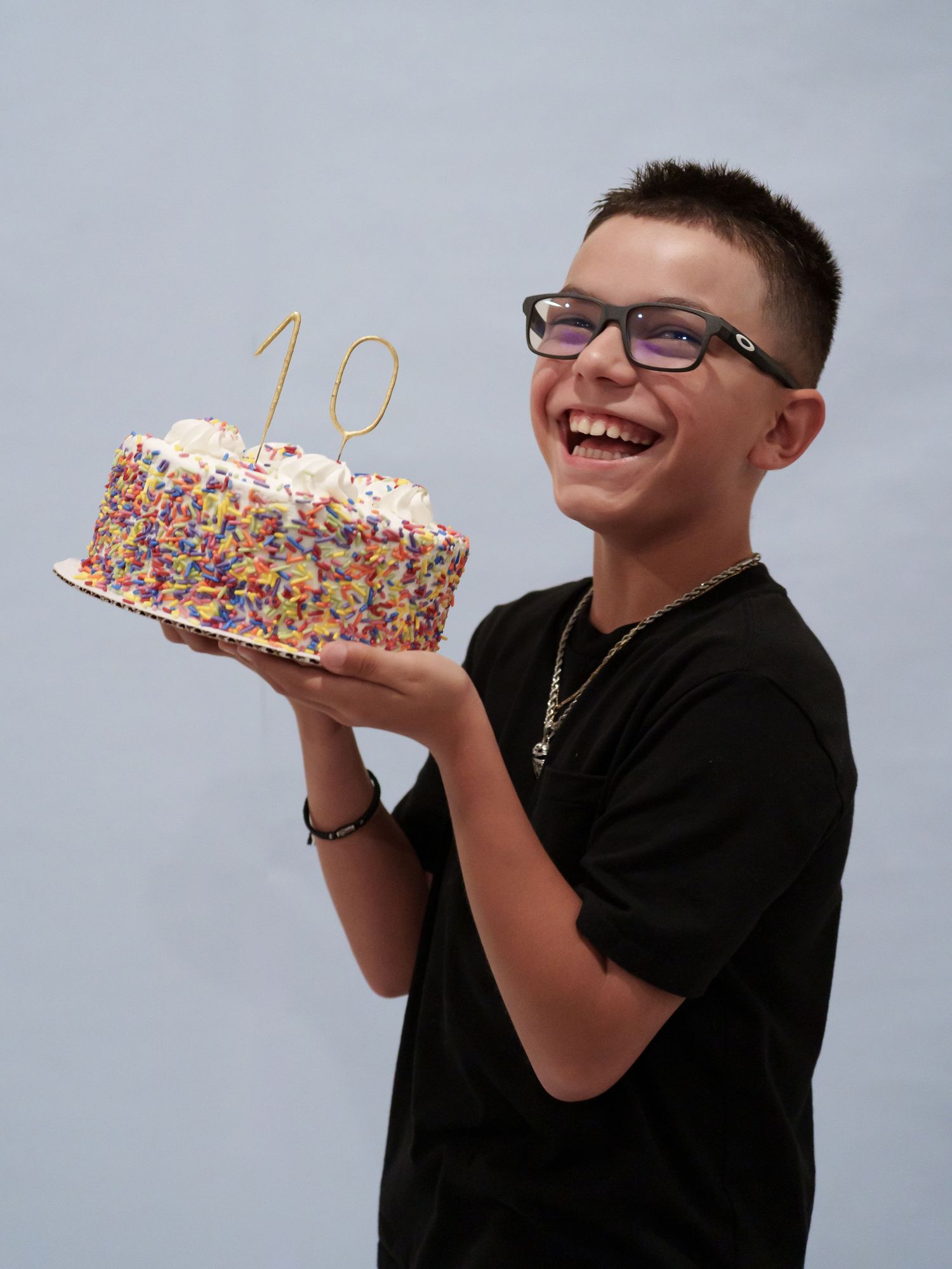 Happy boy in black t-shirt holds colorful sprinkle birthday cake with gold number 10 candle topper.