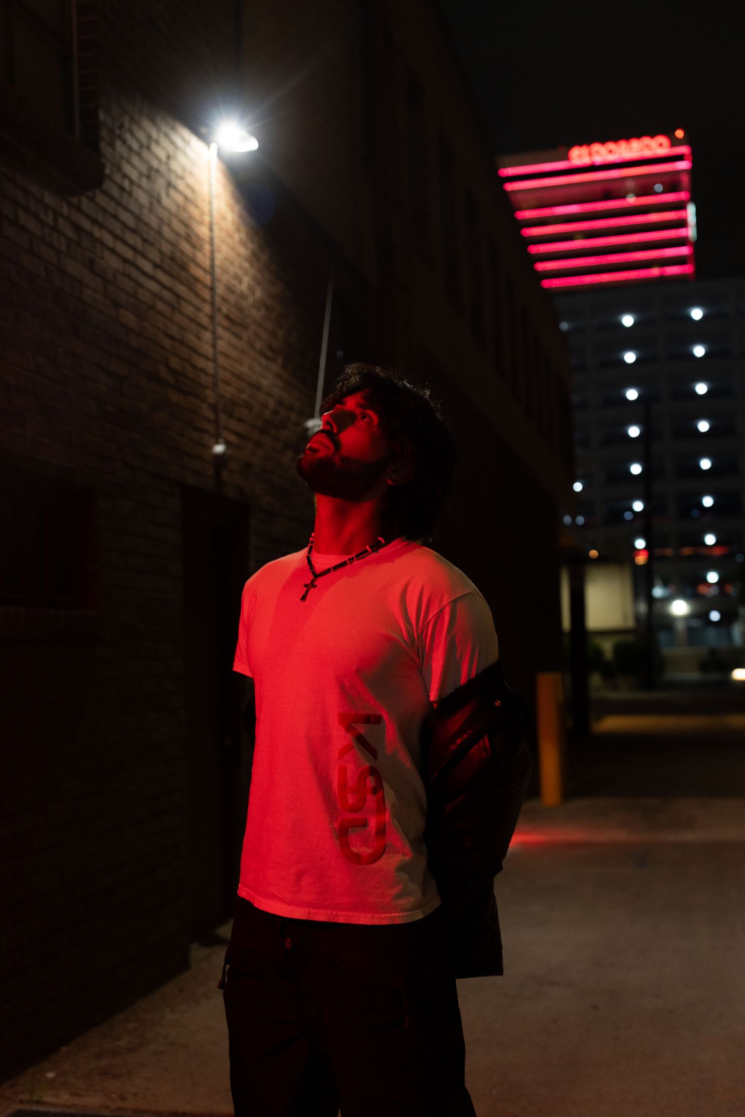 Man in white t-shirt illuminated by red neon light gazes upward in city alley at night.