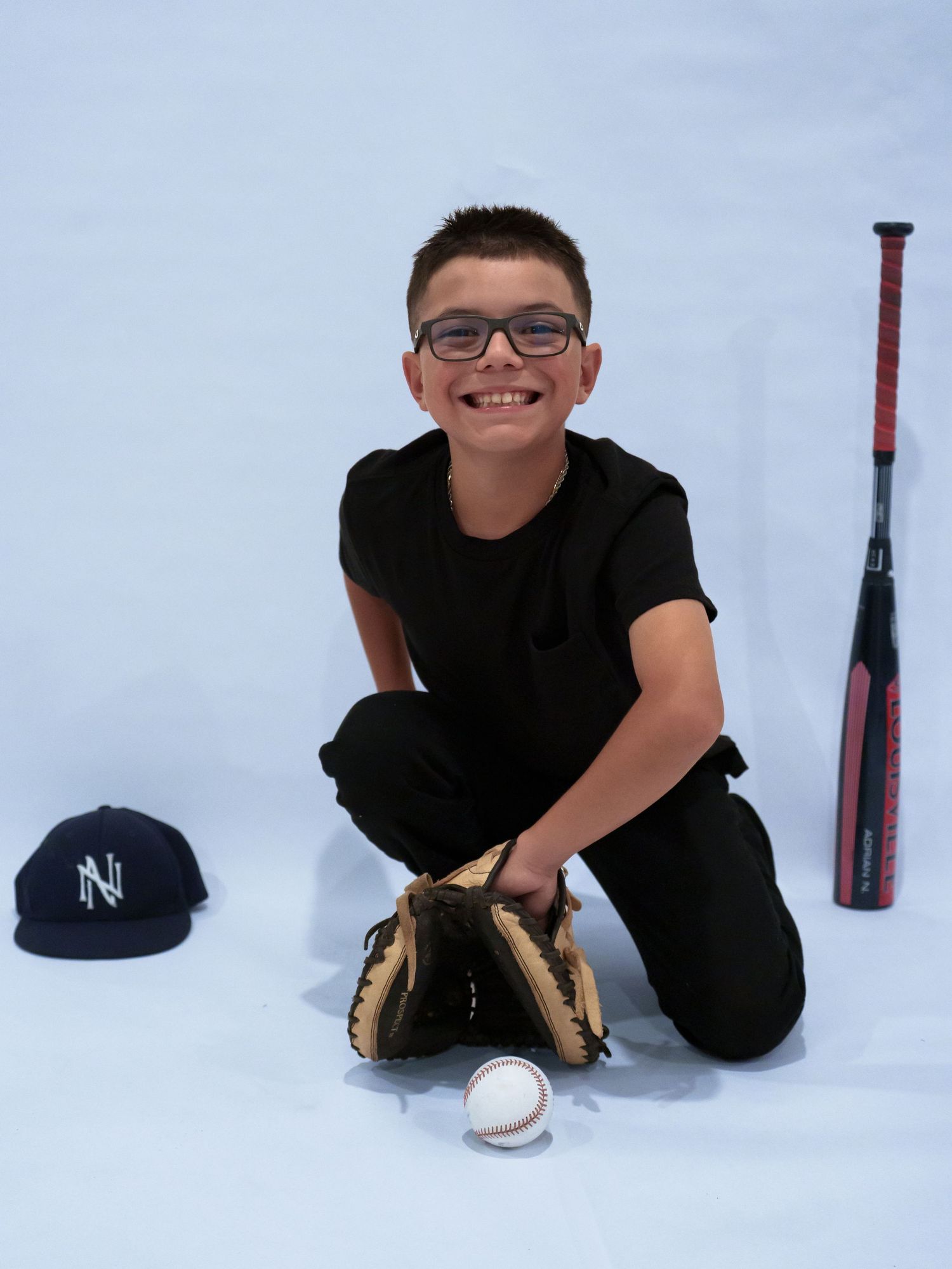 Smiling boy in black outfit crouches with baseball glove, bat, ball, and NY cap on white background.