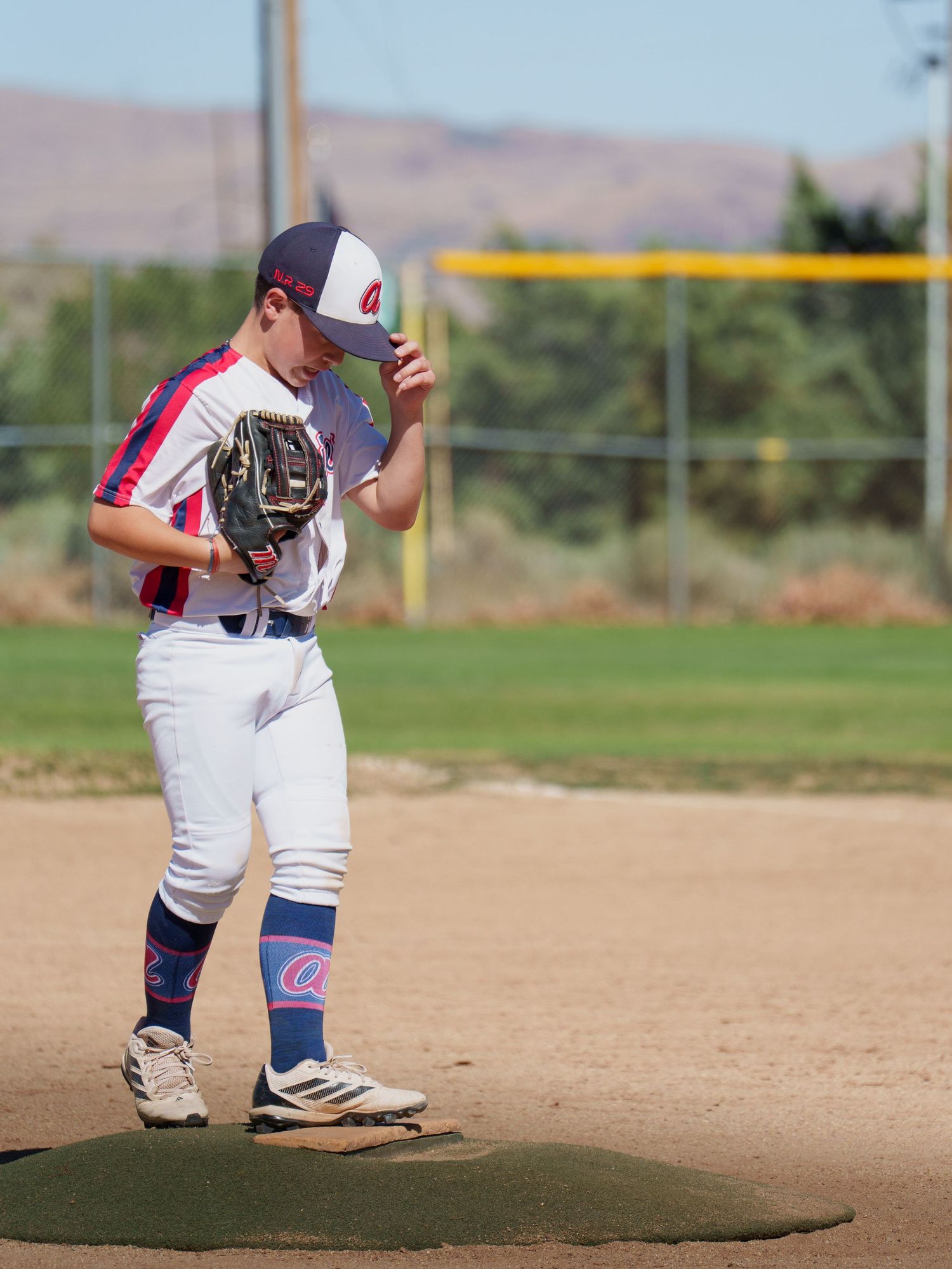 Young baseball pitcher stands on mound adjusting cap wearing white uniform with red and blue accents and baseball glove.