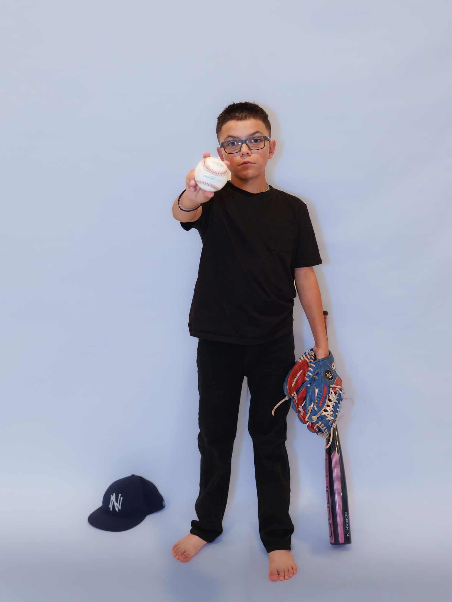 Boy in black outfit holds baseball toward camera with bat, glove, and NY cap on white background.