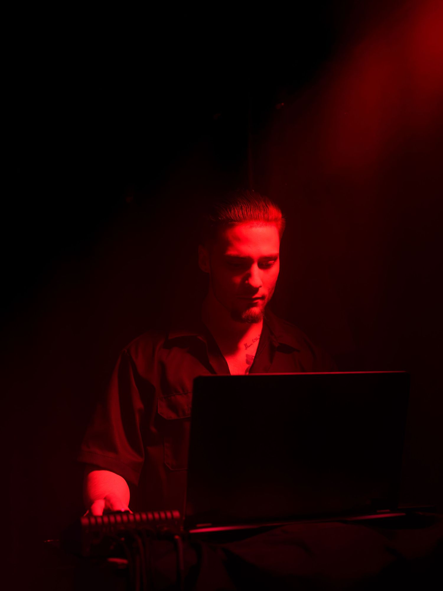 Musician performing with a laptop under dramatic red stage lighting in a dark venue.