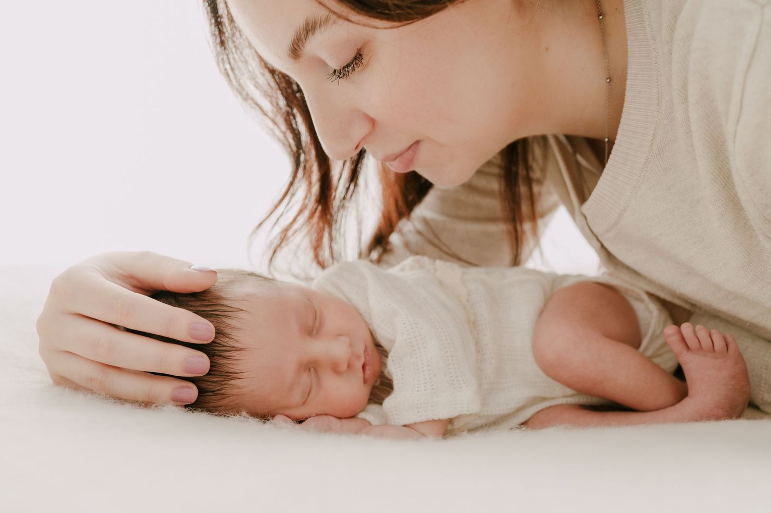 Mother leaning over her peacefully sleeping swaddled newborn baby on a white surface.