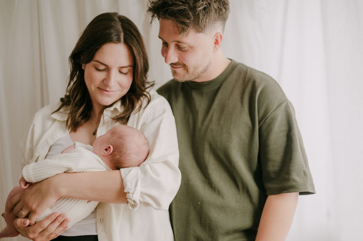 New parents standing together holding sleeping newborn baby in soft white studio setting.