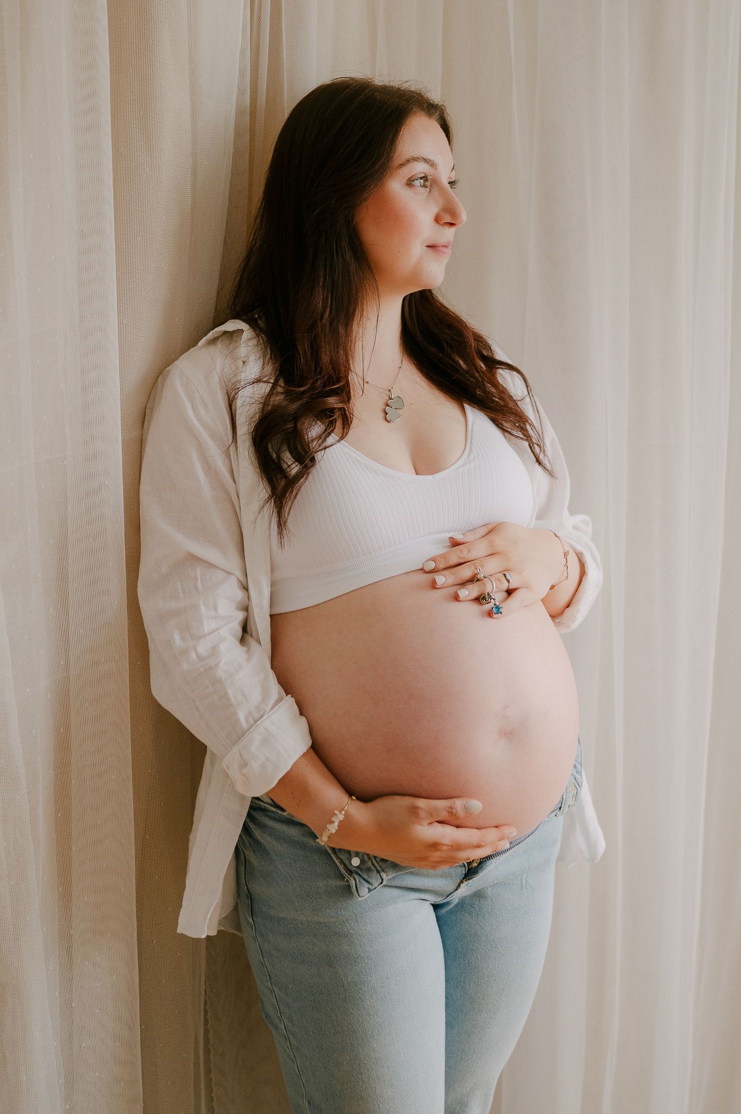 Pregnant woman in white bralette and jeans cradling baby bump against curtain backdrop.