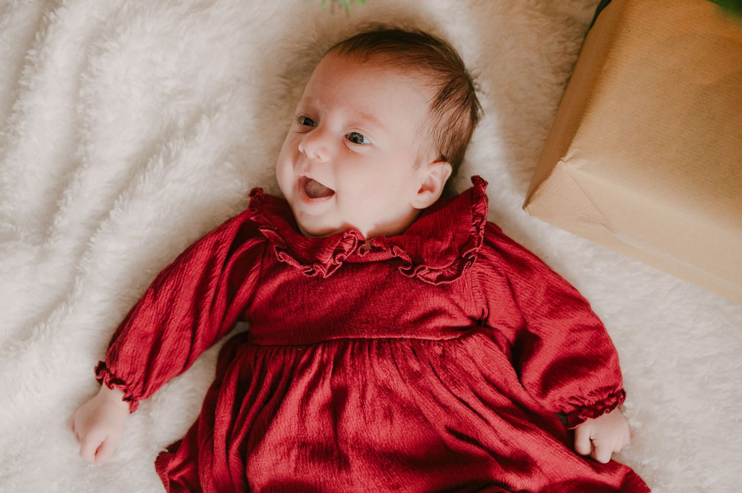 Adorable baby girl in a red ruffled dress lying on a white fluffy blanket during Christmas.