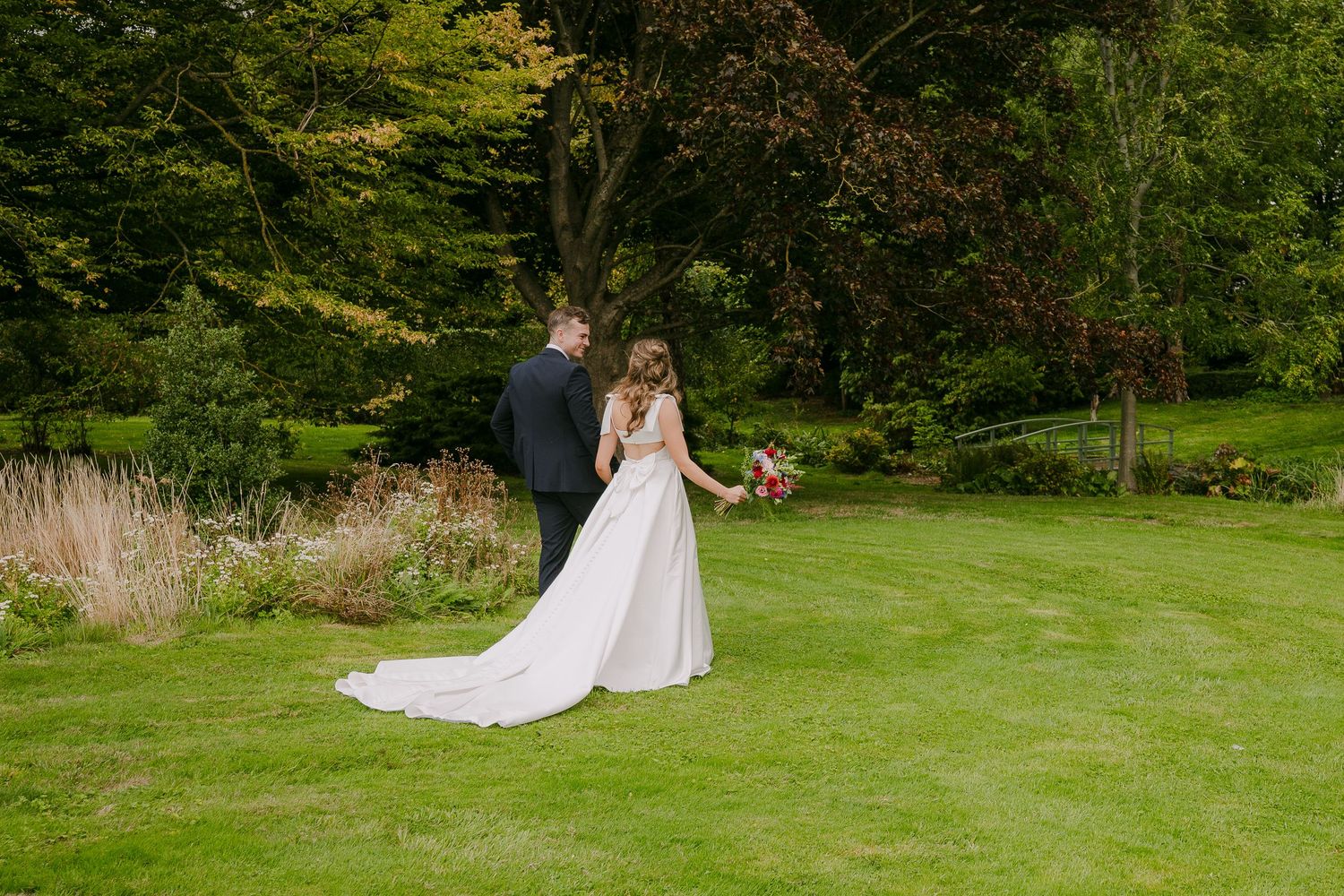 Bride and groom walk hand in hand across a lush green garden with a flowing white wedding gown.