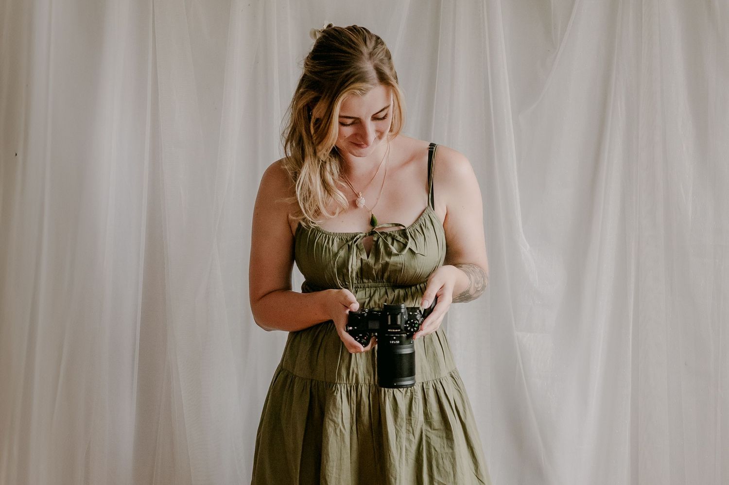Young woman in an olive green dress smiling while holding a camera against a white backdrop.