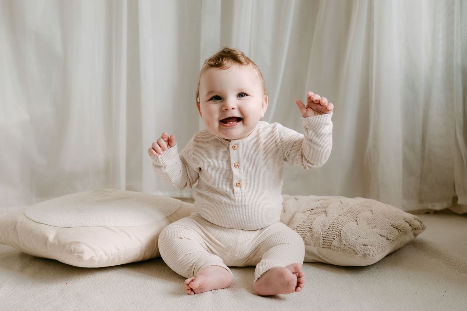 A happy baby in cream outfit sits smiling with arms raised on cushions against a white curtain backdrop.