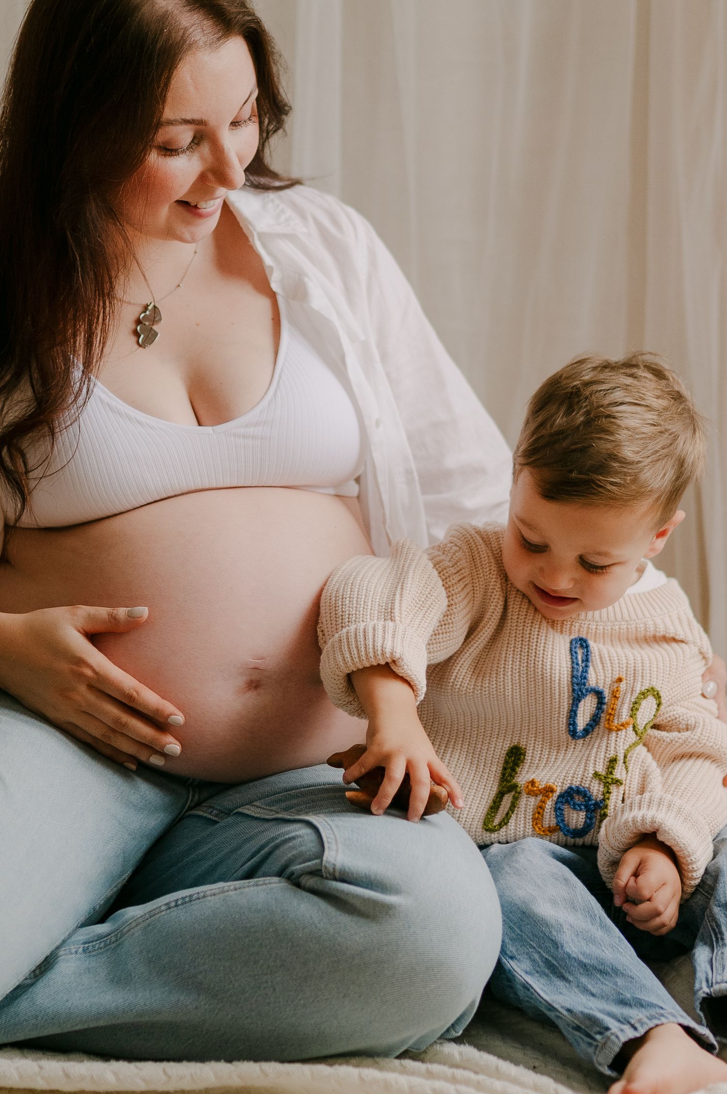 Pregnant mother in white bra and jeans sitting with young son touching her baby bump indoors.