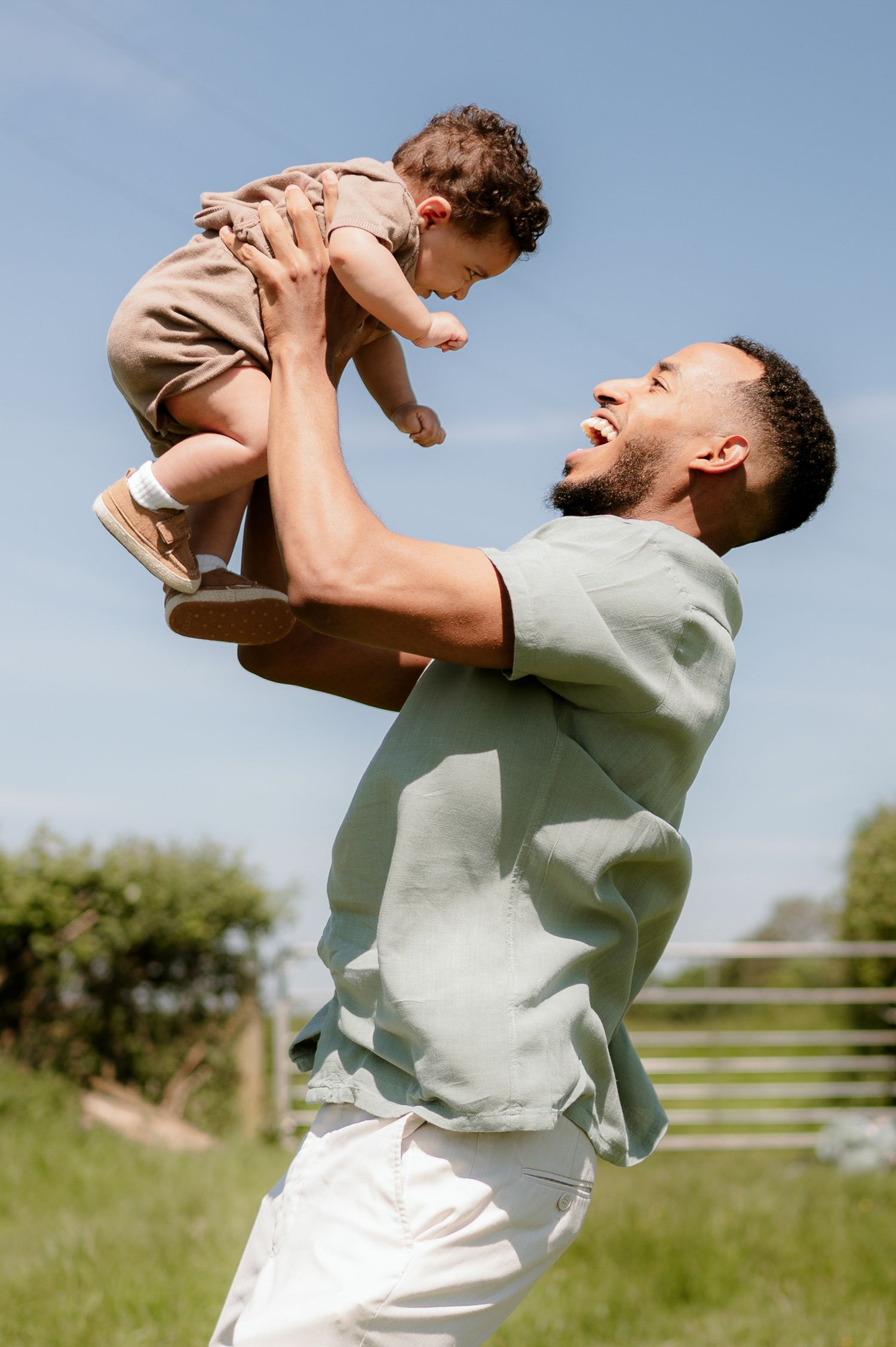 Joyful father lifting laughing baby high in the air outdoors under a blue sky.