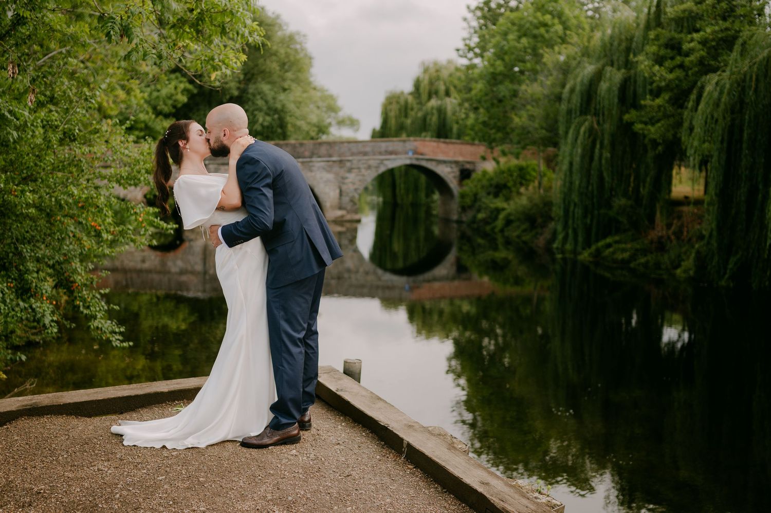 Bride and groom share a romantic kiss by a reflective river with a stone bridge backdrop.