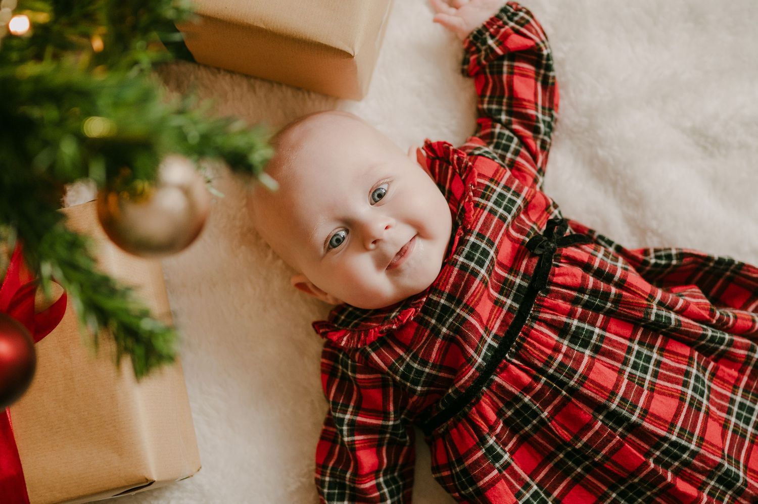 Smiling baby in red tartan Christmas dress lying on white fur blanket beside holiday tree and gifts.