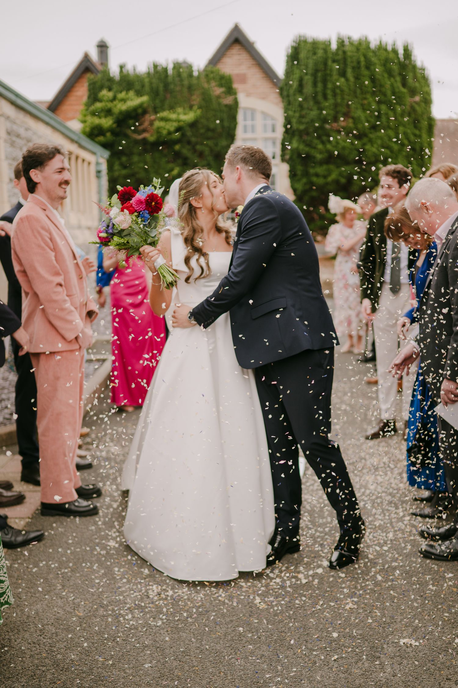 Bride and groom share first kiss surrounded by confetti and smiling guests outside elegant stone venue.