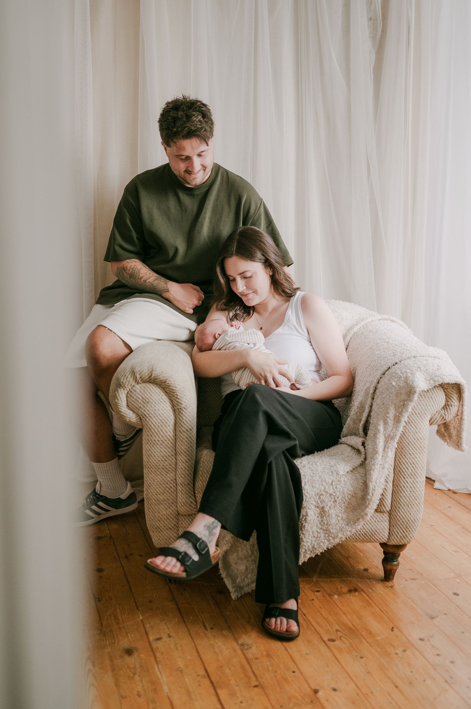 New parents sitting in a cozy armchair together, lovingly gazing at their newborn baby.